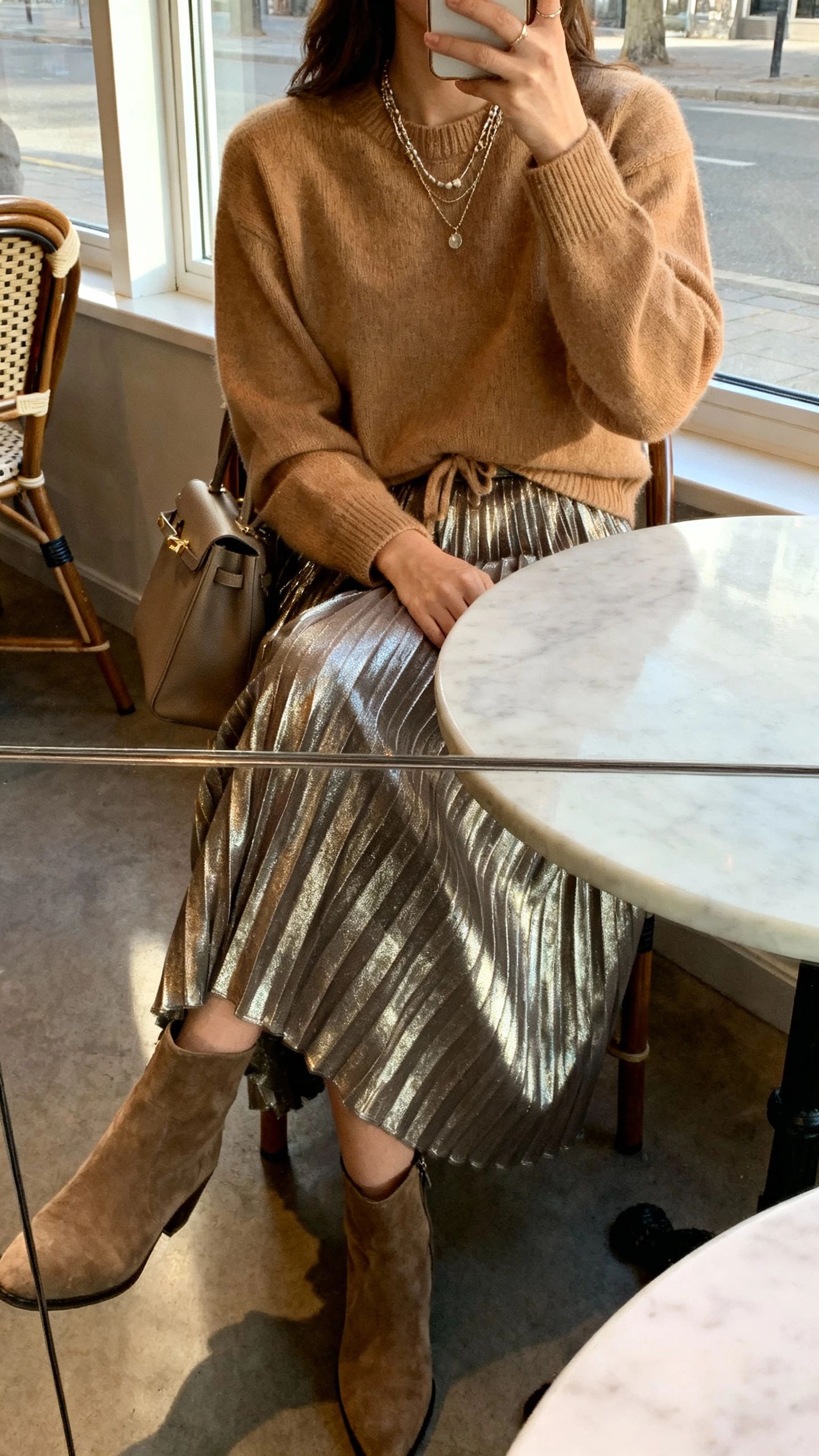 iPhone seated cafe selfie (table reflection) of a woman in metallic pleated midi skirt with a camel cashmere sweater half-tucked, ankle boots, layered necklaces, structured tote, face not visible, morning window light, casual iPhone photo.