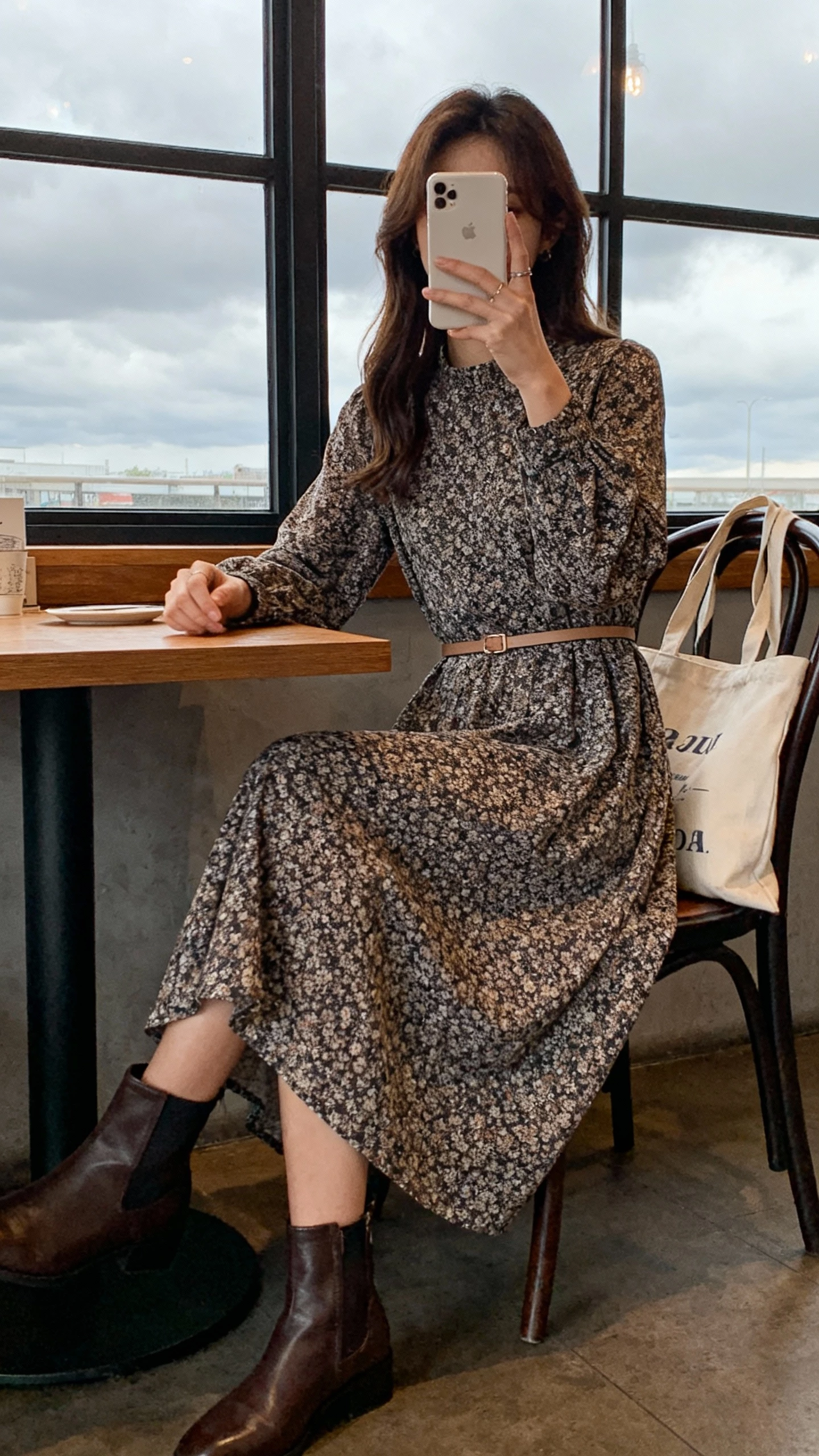 iPhone seated selfie at a cafe table showing a woman in a printed long-sleeve midi dress, ankle boots, thin belt at waist, and a tote bag on chair, face not visible, window light on a cloudy day, natural iPhone photo.