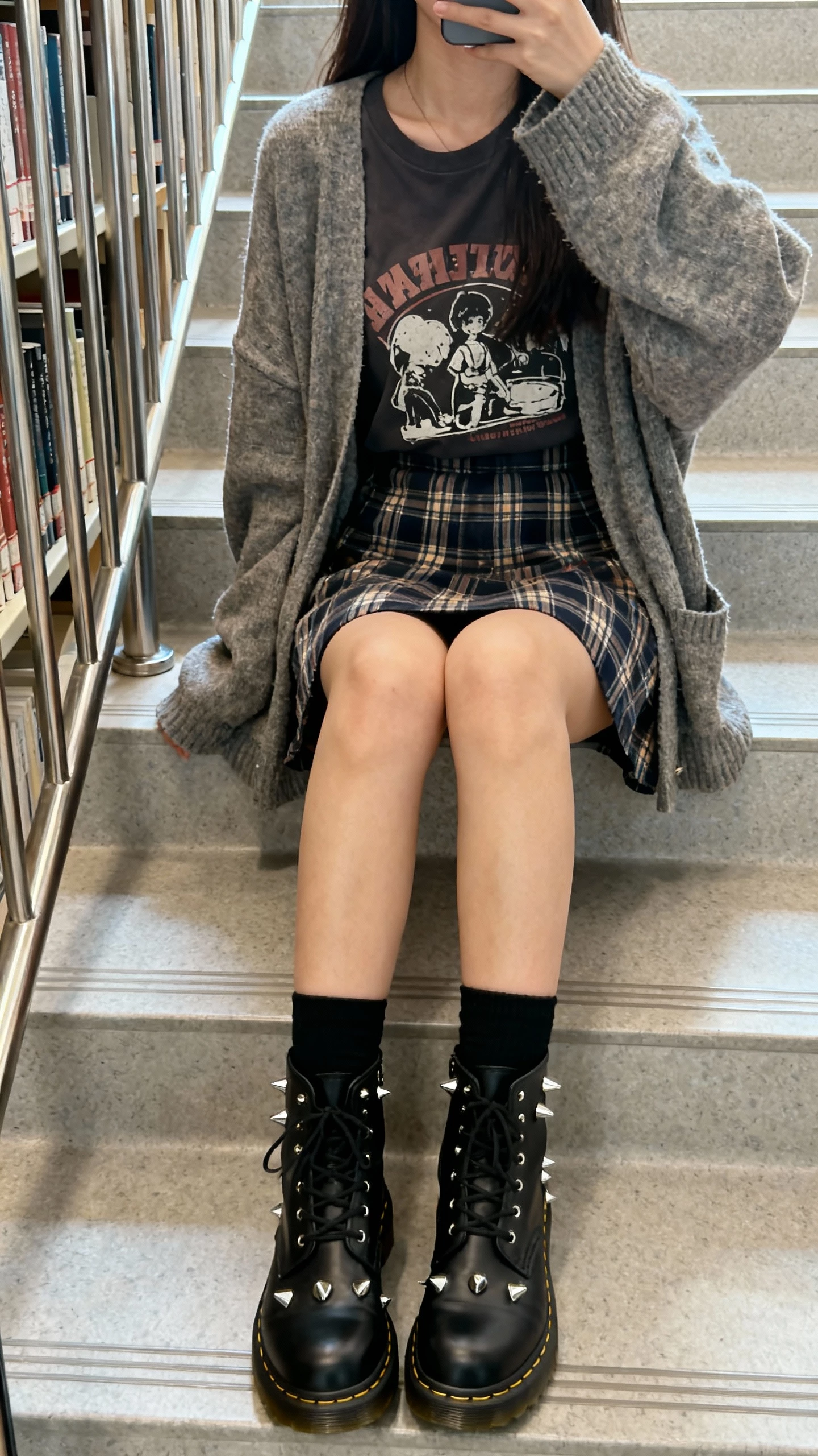 iPhone seated selfie cropped at neck of a woman wearing a plaid skirt, graphic tee, slouchy cardigan, black Doc Martens, black socks, silver studs, face not visible, sitting on a library stairwell, soft daylight, casual iPhone photo.