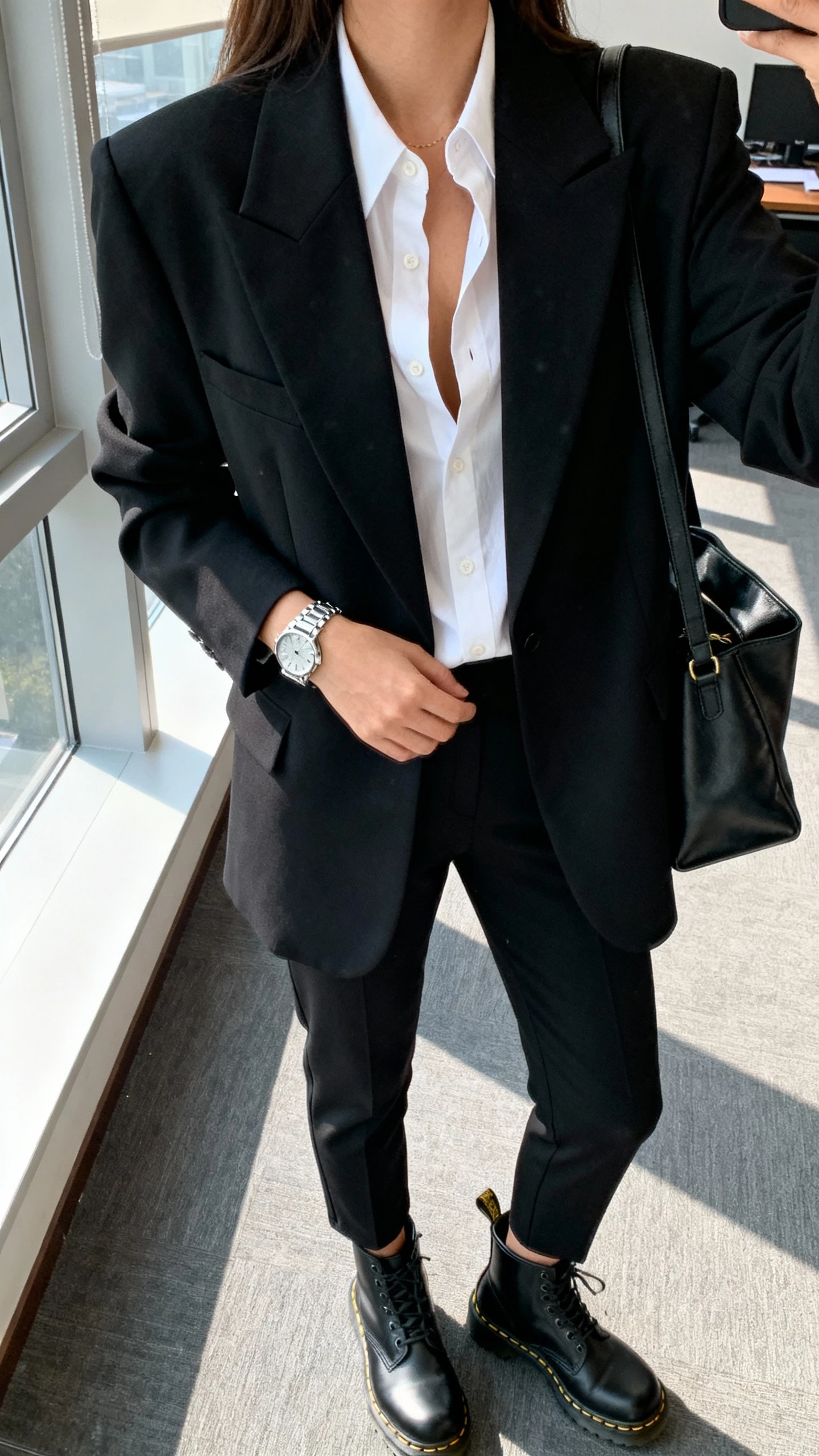 iPhone selfie cropped at neck showing a woman in a modern black suit (boxy blazer and tapered trousers) with a crisp white button-up slightly unbuttoned, black Doc Martens, silver watch, and structured tote, face not visible, office setting by a window with natural morning light, natural iPhone photo quality.