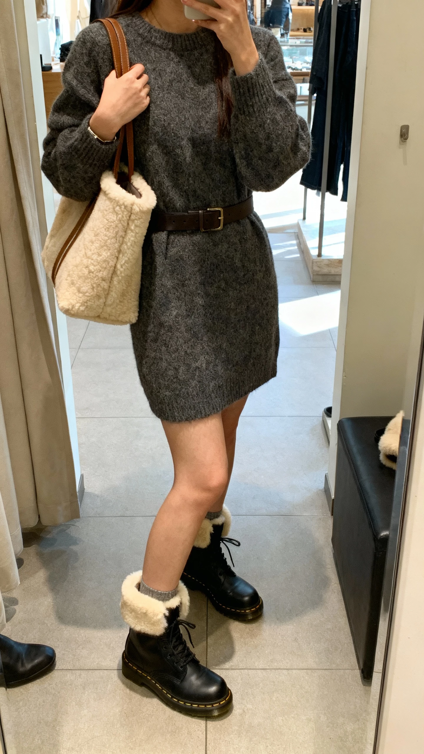 iPhone selfie cropped at neck showing a woman in a sweater dress cinched with a belt, shearling tote and shearling-trim accessories, Doc Martens, face not visible, boutique fitting-room mirror, indoor lighting, natural iPhone quality.