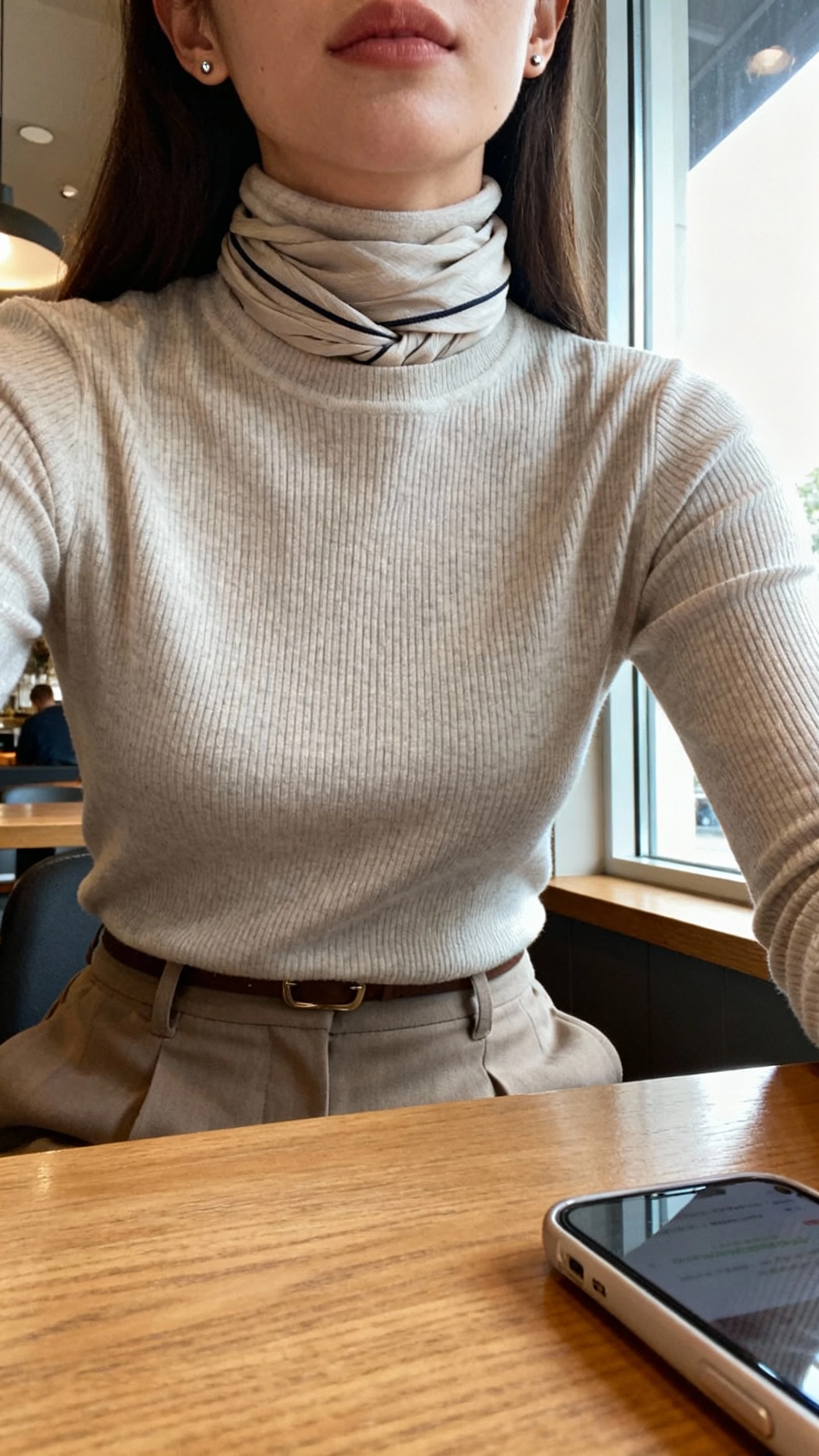 iPhone selfie cropped at neck showing a woman wearing a fine ribbed turtleneck with a thin scarf tucked neatly inside the collar for a sleek layered illusion, high-waist trousers, minimal stud earrings, face not visible, desk-side at a cafe, window light, natural iPhone aesthetic.