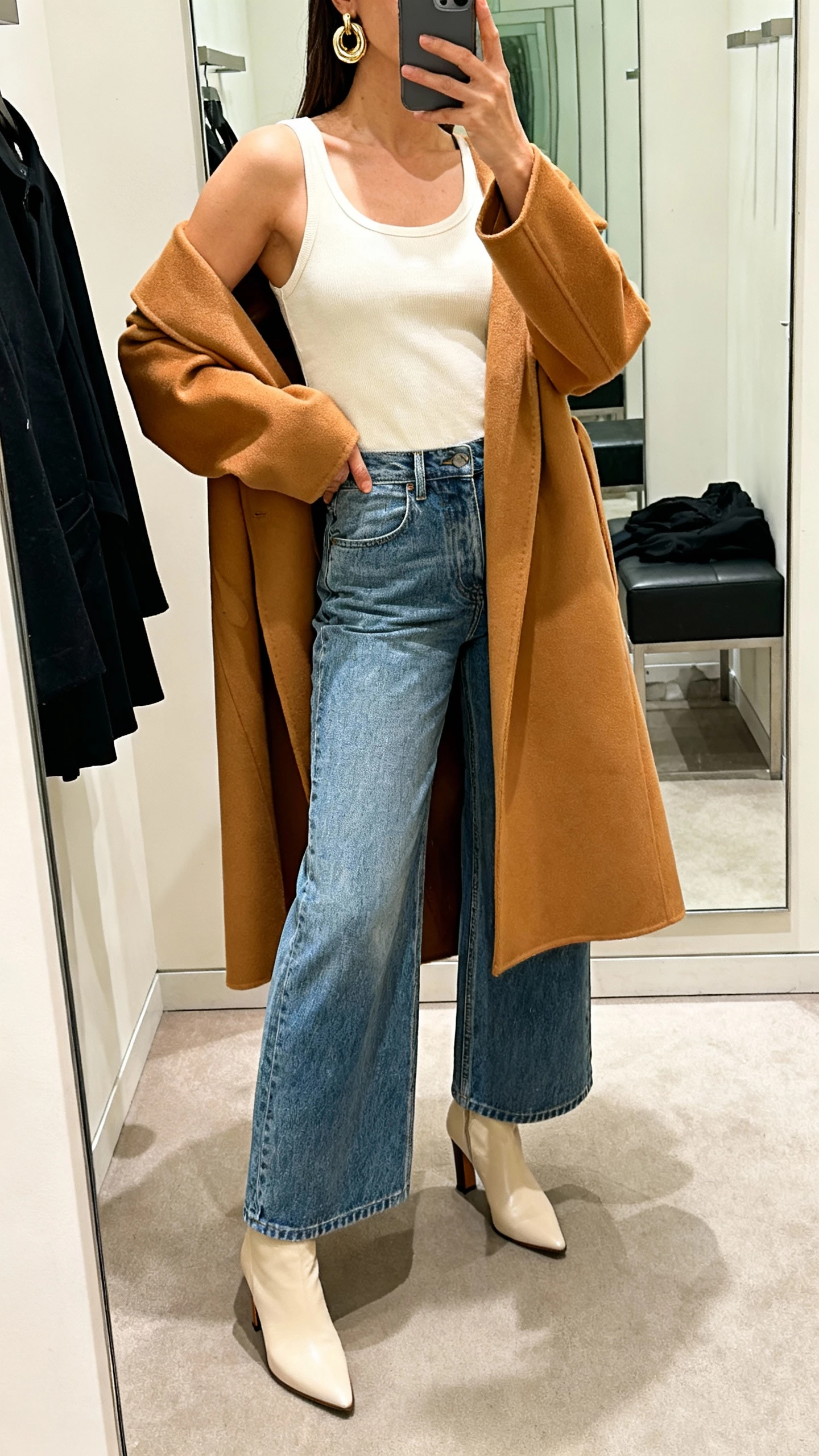 iPhone selfie cropped at neck showing a woman wearing wide leg jeans, a sleek cream minimalist tank, a bold camel statement coat draped over, heeled ankle boots and chunky earrings, face not visible, boutique fitting-room mirror, warm indoor lighting, natural iPhone photo quality.
