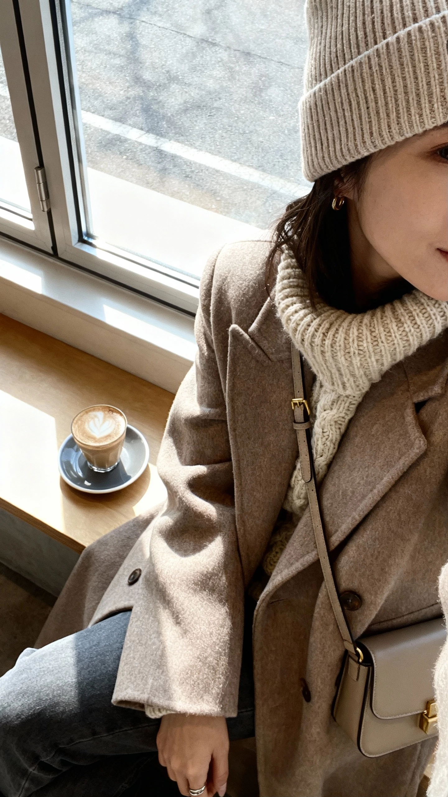 iPhone selfie cropped at shoulders of a woman in a ribbed chic beanie, chunky knit sweater, long coat, small crossbody bag, face not visible, cozy coffee shop window seat with latte, soft afternoon light, natural iPhone aesthetic.