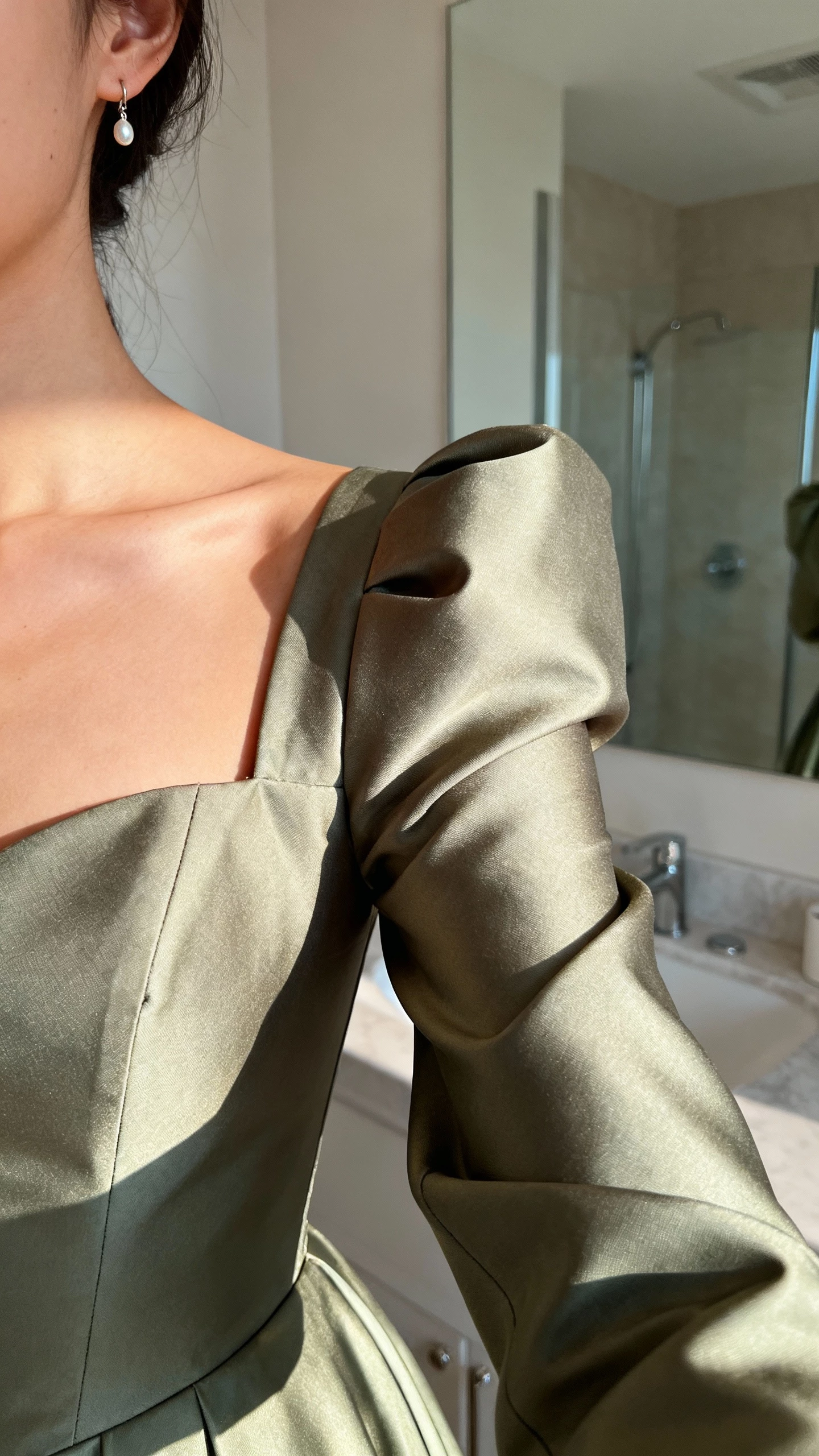 iPhone selfie cropped at shoulders of a woman in a gown with adjusted straps and tailored sleeves that sculpt the shoulders and frame the collarbone; small drop earrings, face not visible, bathroom mirror, soft indoor lighting, natural iPhone photo quality