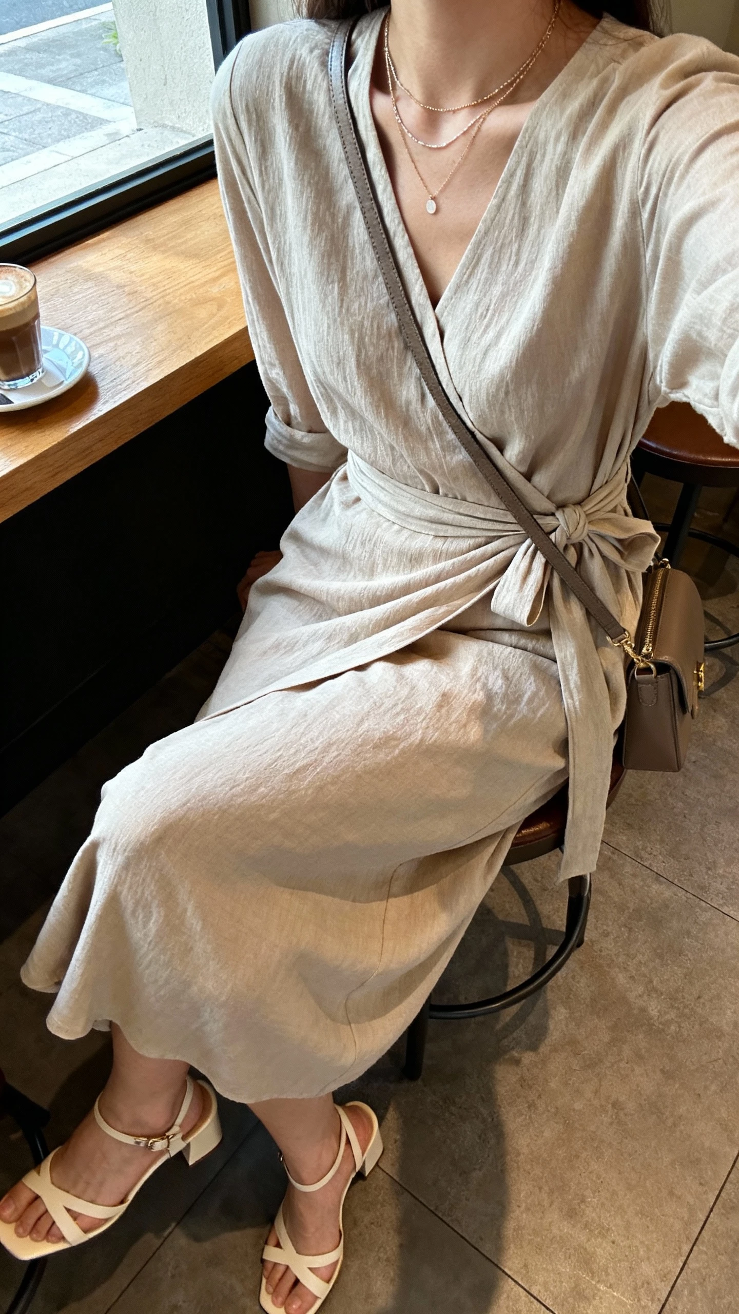 iPhone selfie cropped at shoulders of a woman in a wrap-style midi dress tied at the waist, heeled sandals, layered delicate necklaces, and a crossbody bag, face not visible, coffee shop seating by window, soft morning light, natural iPhone aesthetic.