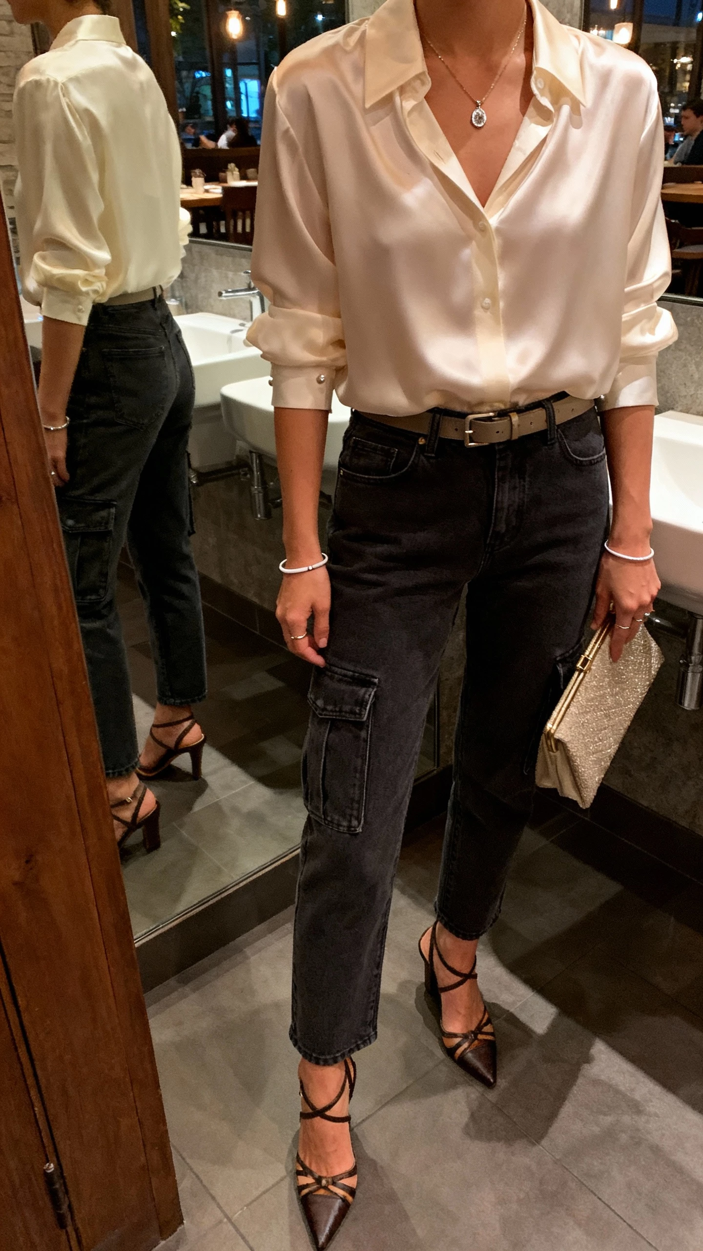 iPhone selfie cropped at shoulders of a woman in an ivory silk blouse with tailored dark-rinse cargo jeans, strappy heels, refined clutch, delicate pendant necklace, thin bangles, subtle studs, and a slim belt, face not visible, restaurant restroom mirror, warm evening lighting, natural iPhone aesthetic.
