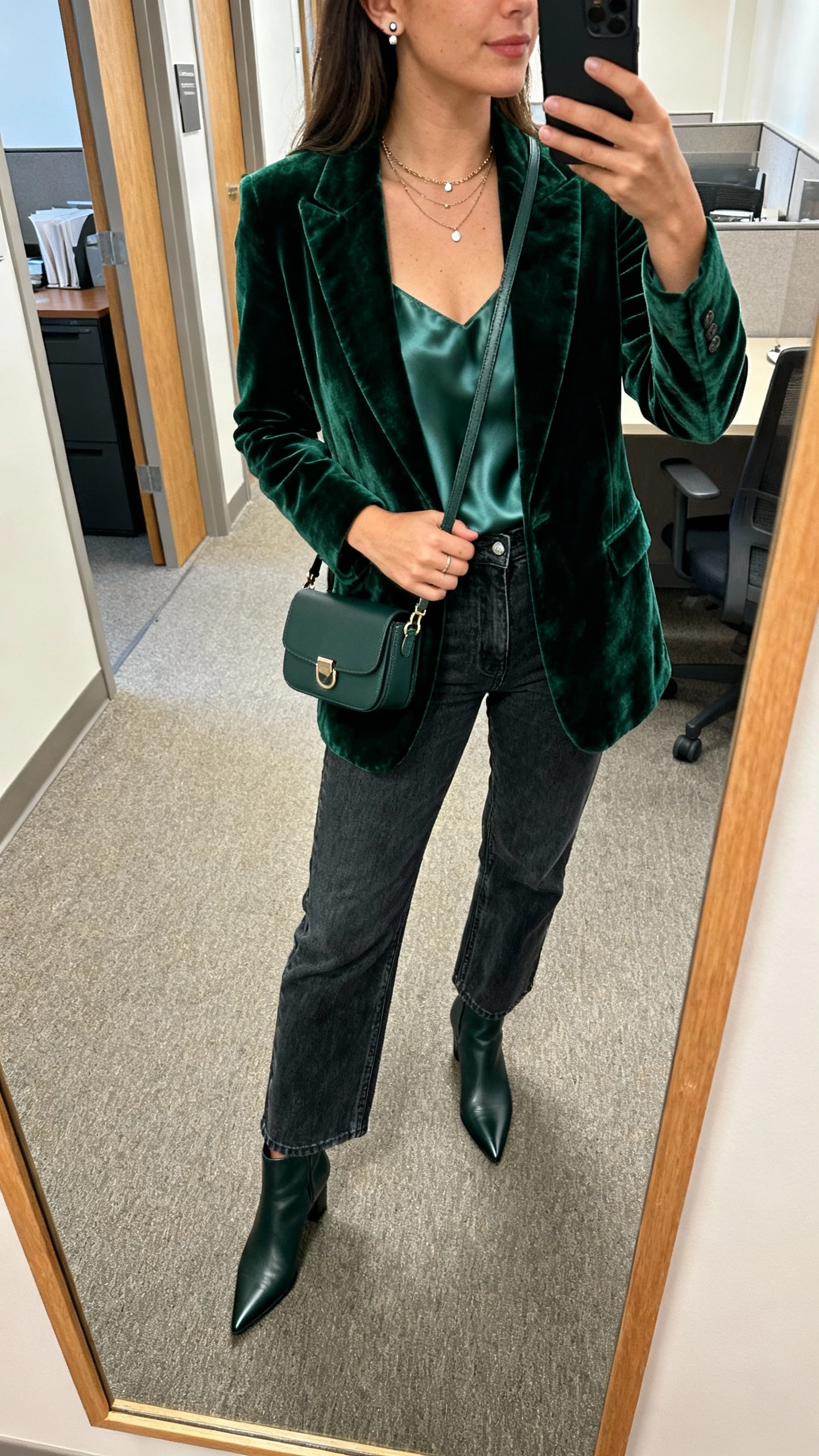 iPhone selfie cropped at shoulders of a woman wearing a deep green velvet blazer over a silky camisole, dark-wash straight-leg jeans, pointed-toe ankle boots, small crossbody with subtle hardware, layered necklaces and stud earrings, office hallway mirror, morning light, face not visible, natural iPhone photo quality.