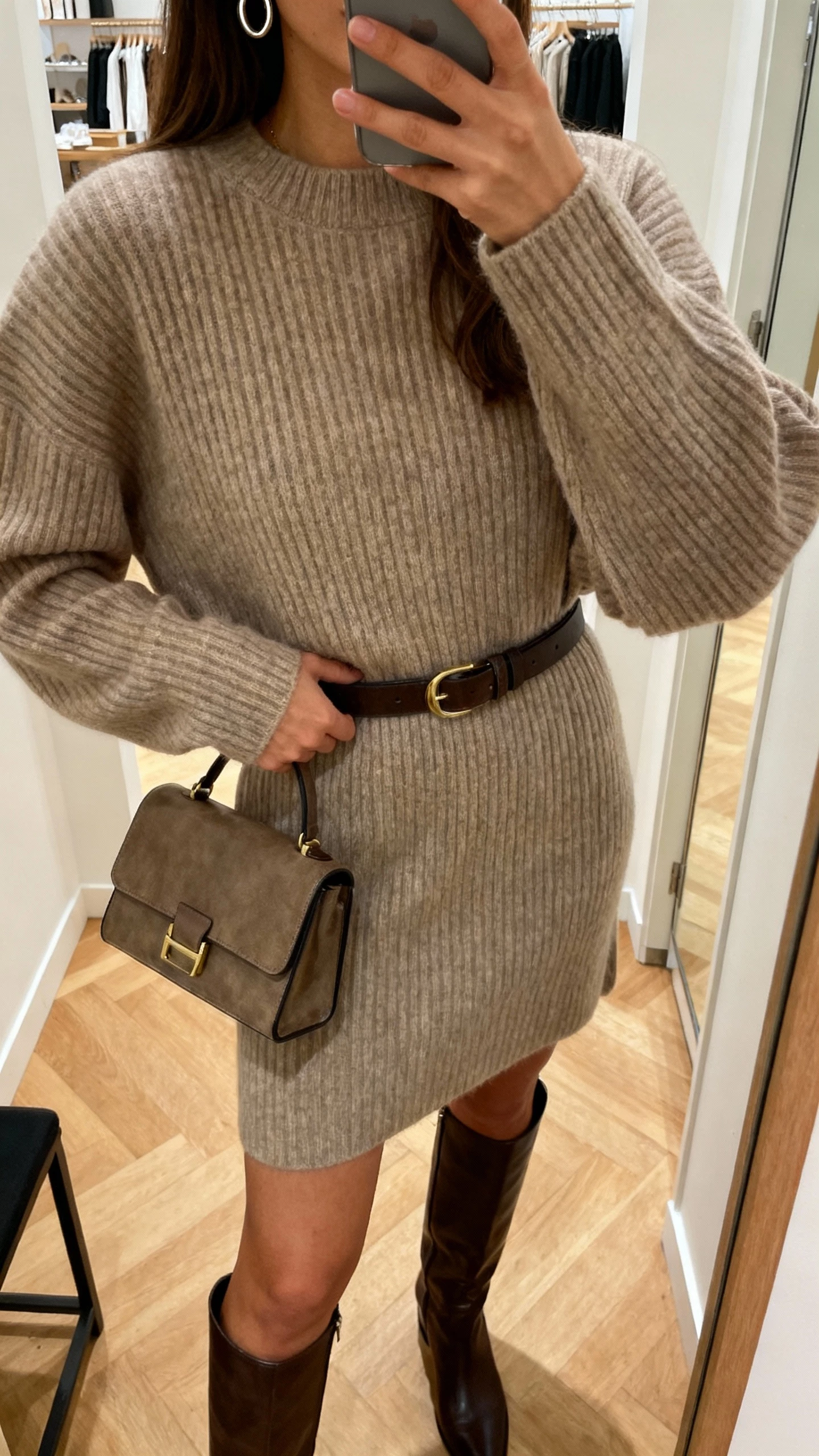 iPhone selfie cropped at shoulders showing a woman in a ribbed sweater dress with knee-high boots, belt at waist, small structured handbag, simple hoop earrings, face not visible, boutique dressing-room mirror, warm indoor lighting, natural iPhone photo quality.
