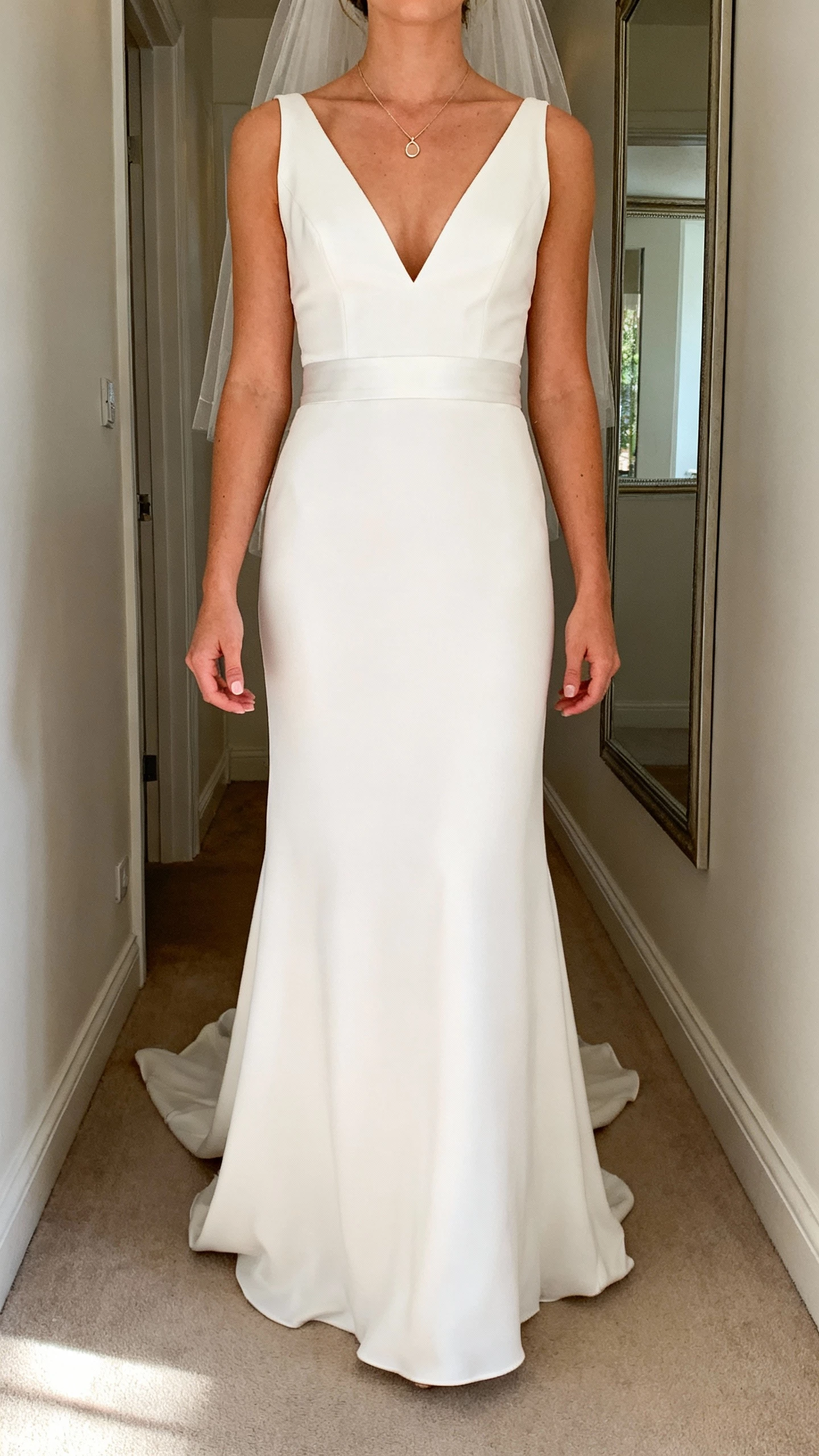 iPhone selfie cropped at shoulders showing a woman in a chic V-neck wedding gown with a slimming waist, subtle plunge, delicate pendant necklace, minimal veil, face not visible, hallway mirror, morning light, natural iPhone aesthetic.