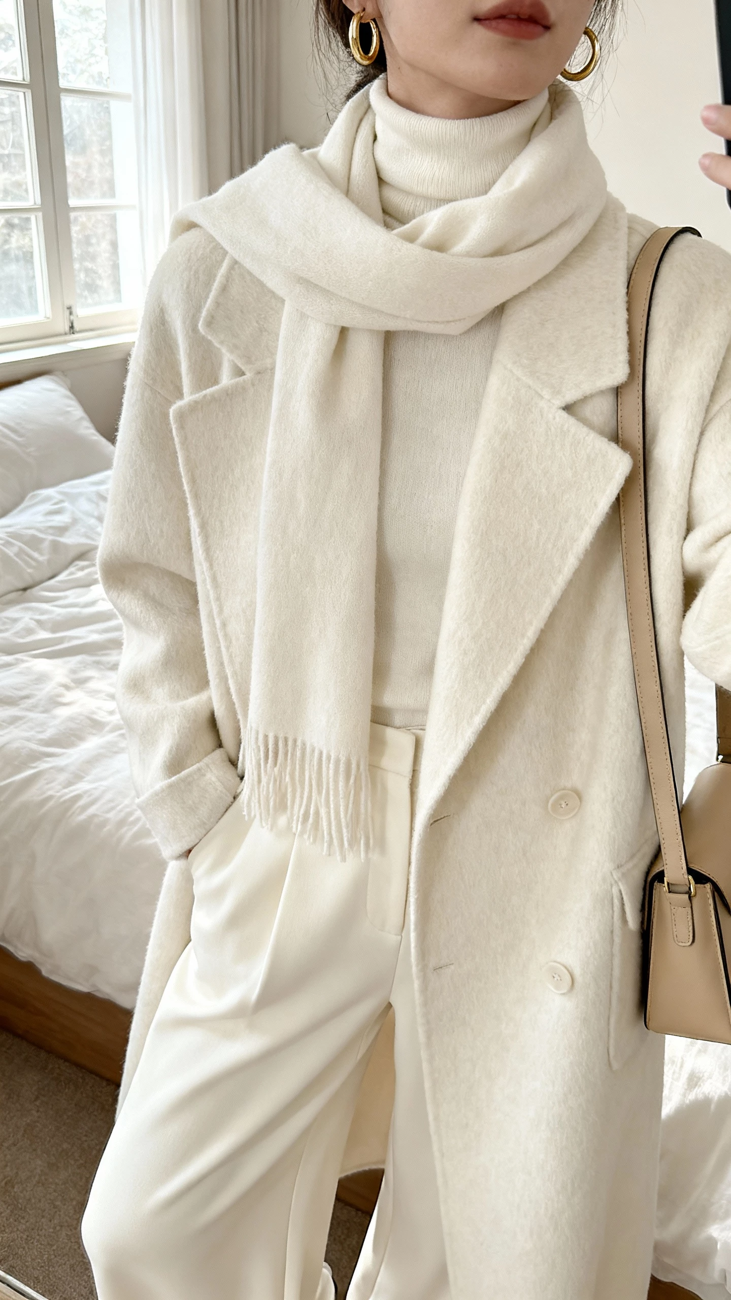 iPhone selfie cropped at shoulders showing a woman in monochrome cream layers: cream turtleneck, cream wool coat, cream wide-leg trousers, cream scarf, beige shoulder bag, gold hoops, face not visible, soft window light in a cozy bedroom, natural iPhone photo quality.