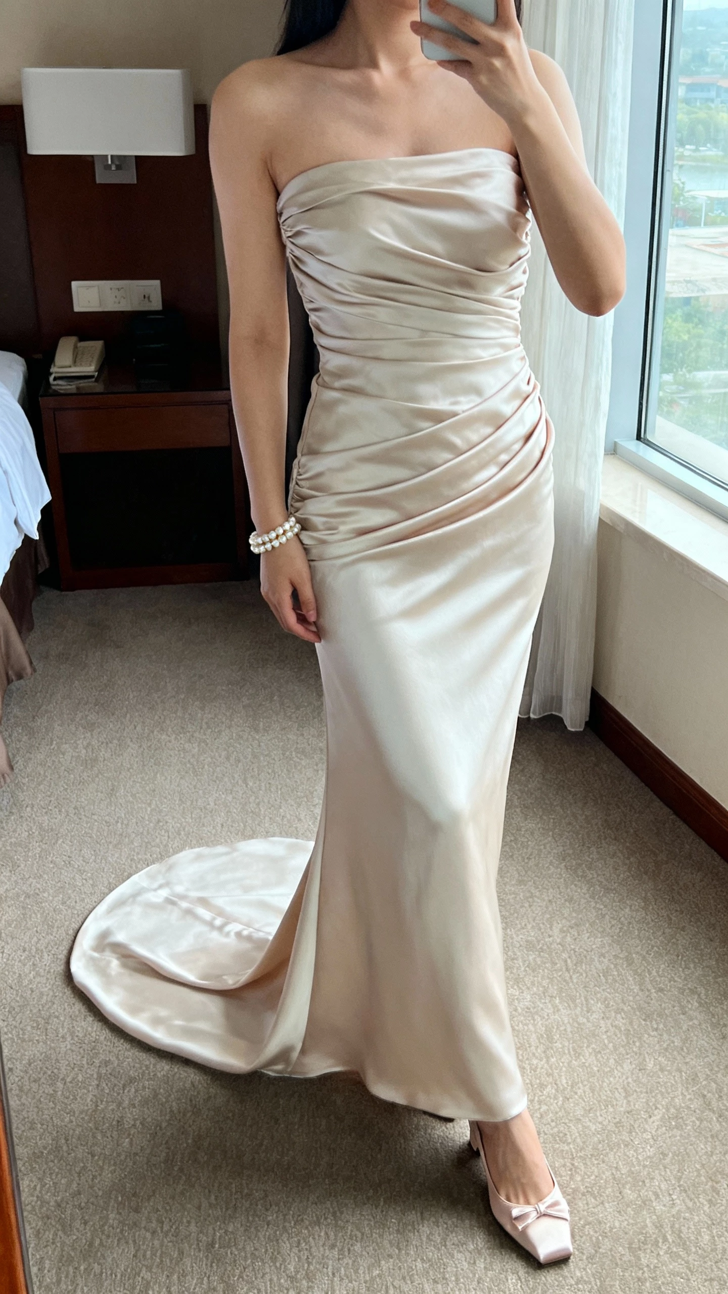iPhone selfie cropped at shoulders showing a woman in a strapless draped silk column gown with gentle ruching and a smooth train, pearl bracelet and satin slingback heels, face not visible, hotel room window light, natural iPhone photo quality.
