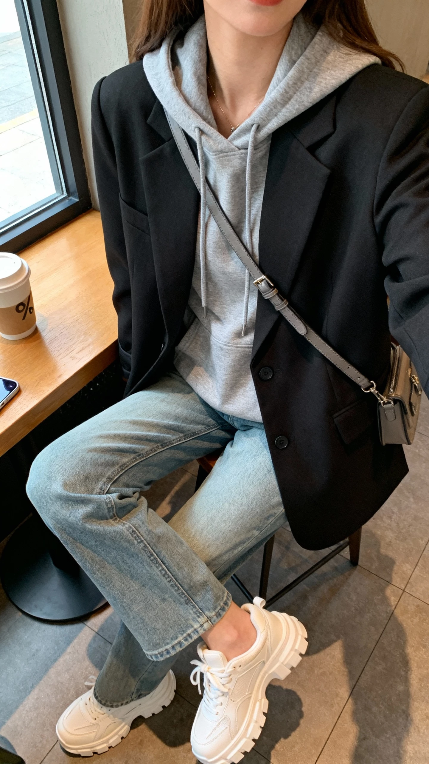 iPhone selfie cropped at shoulders showing a woman layering a gray hoodie under a black blazer with light blue jeans, chunky white sneakers, crossbody bag, face not visible, coffee shop table shot with natural window light, natural iPhone photo quality