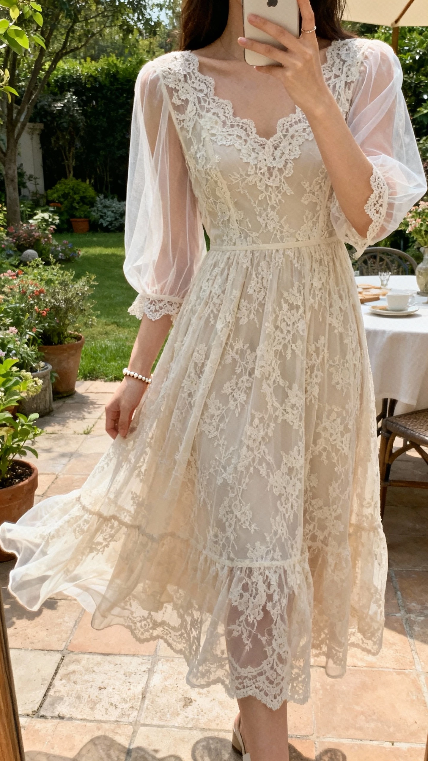 iPhone selfie cropped at shoulders showing a woman in a romantic A-line lace gown with sheer sleeves and airy movement, delicate lace details, dainty bracelet, face not visible, garden patio setting, gentle afternoon light, natural iPhone photo quality