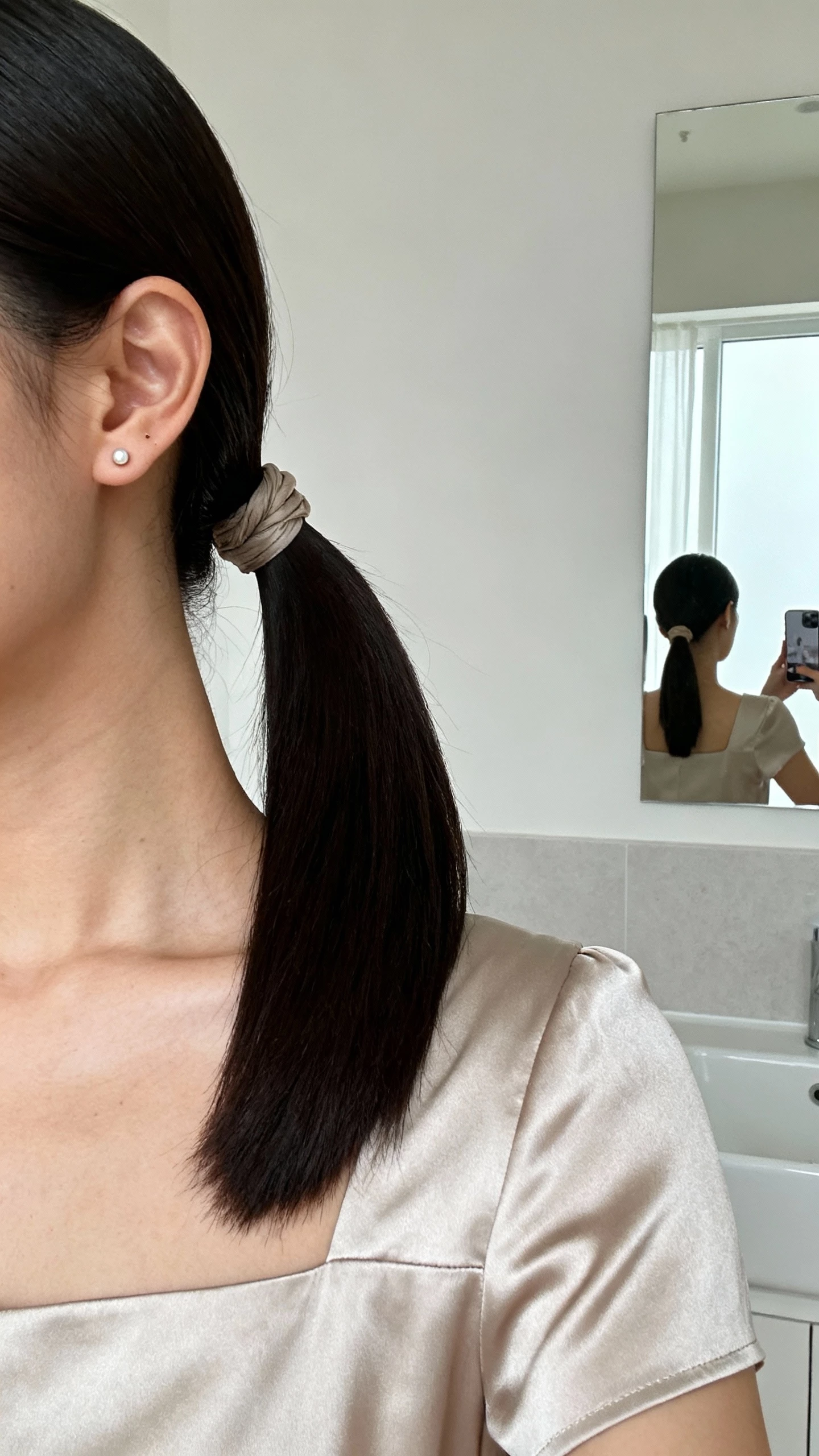 iPhone selfie cropped at shoulders showing a woman with a sleek low ponytail with a wrapped base, minimalist stud earrings, square-neck silk dress, face not visible, bathroom mirror, bright indoor lighting, natural iPhone photo quality.