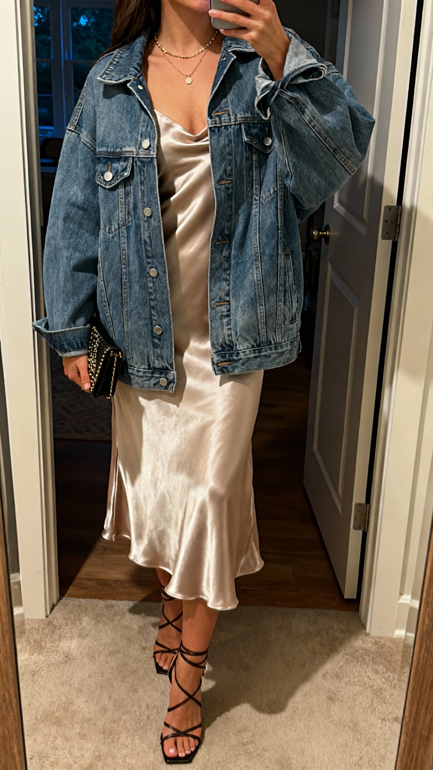 iPhone selfie cropped at shoulders showing a woman in an oversized denim jacket over a satin slip dress, delicate necklace, strappy heels, small clutch, face not visible, evening apartment doorway mirror, warm indoor light, natural iPhone photo quality