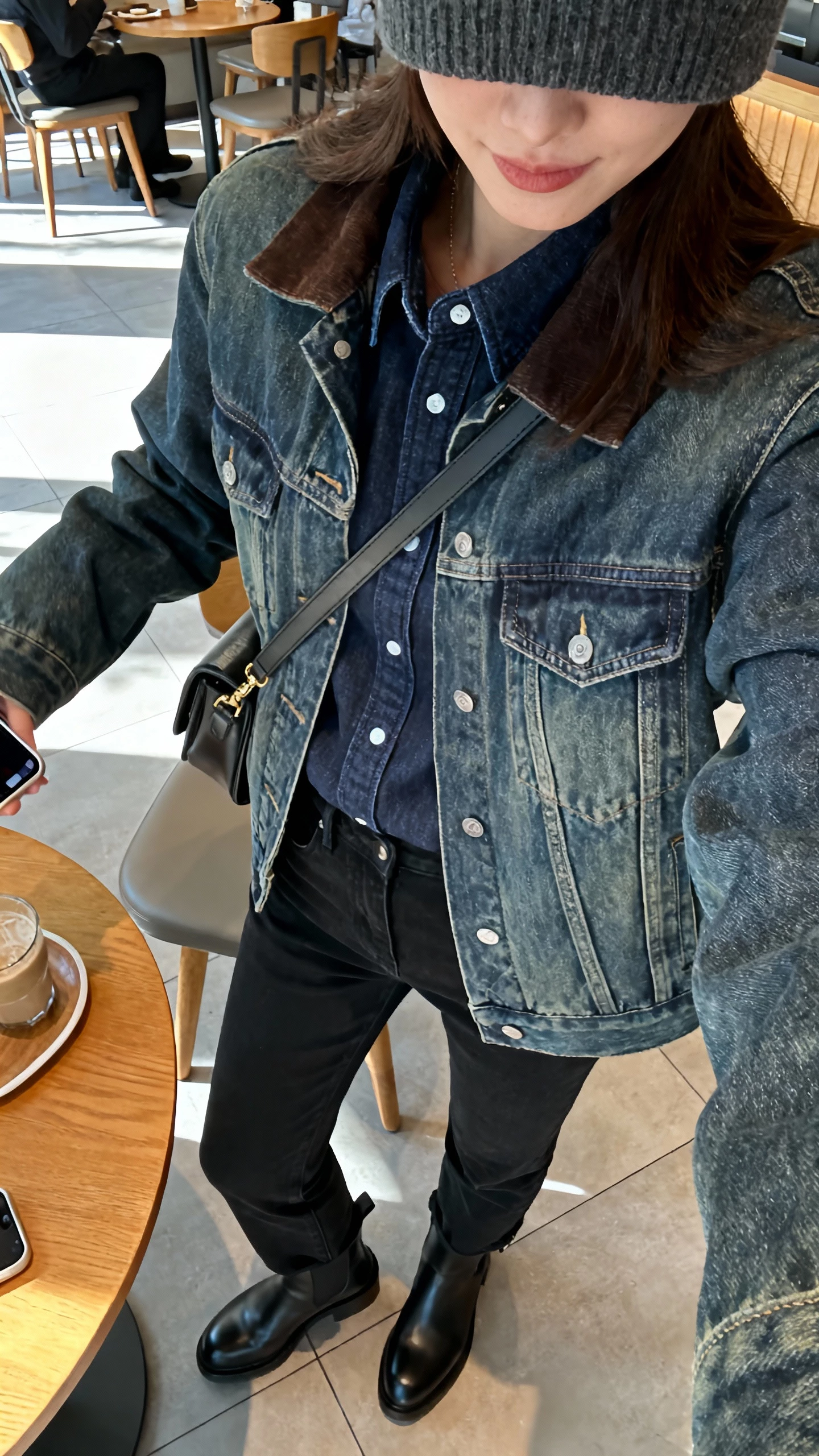 iPhone selfie cropped at shoulders showing a woman in winter denim-on-denim: dark-wash denim shirt layered under a medium-wash denim jacket, black straight-leg jeans, black Chelsea boots, beanie, crossbody bag, face not visible, cafe table setting with natural light, casual iPhone photo.