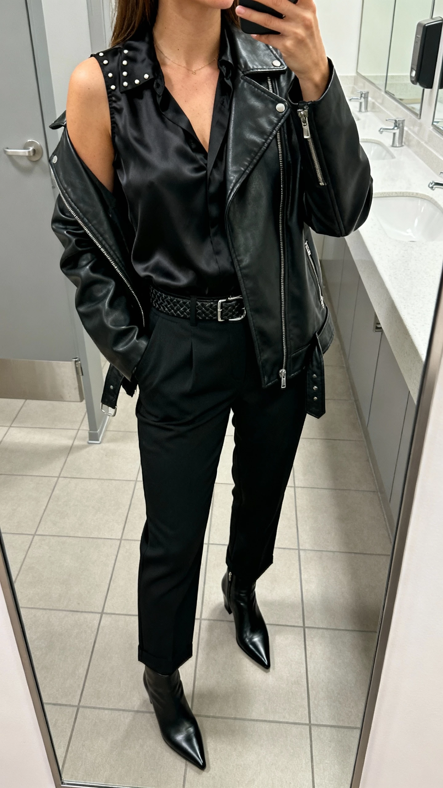 iPhone selfie cropped at shoulders showing a woman in an all-black look: satin blouse, high-waisted trousers, leather jacket, pointed ankle boots, subtle textured belt and minimal studs, face not visible, office restroom mirror with bright indoor light, natural iPhone photo.