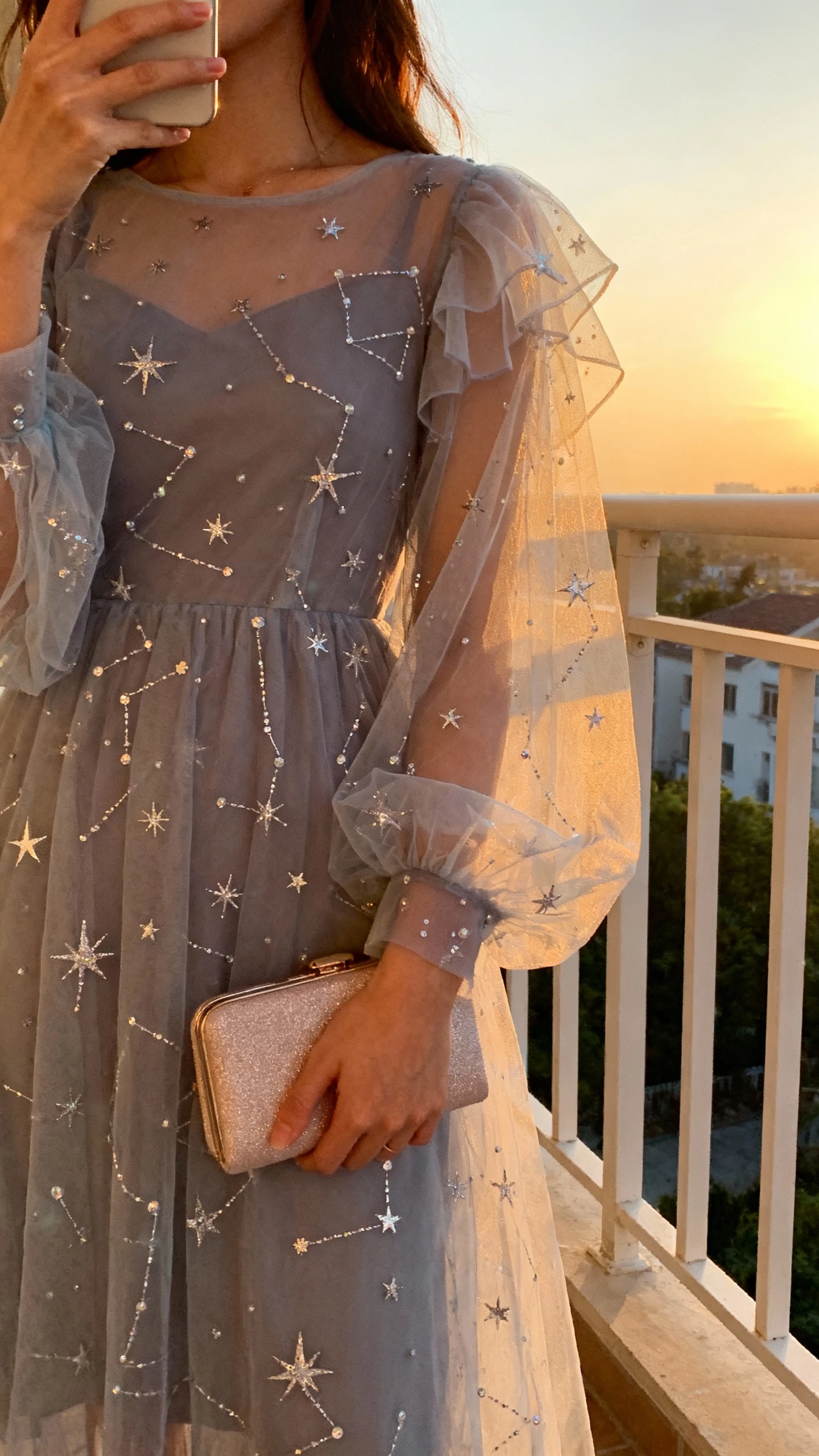 iPhone selfie cropped at shoulders showing a woman in starry tulle with constellation beading, sheer layered sleeves, subtle shimmer clutch, face not visible, balcony at dusk, natural ambient light, iPhone photo