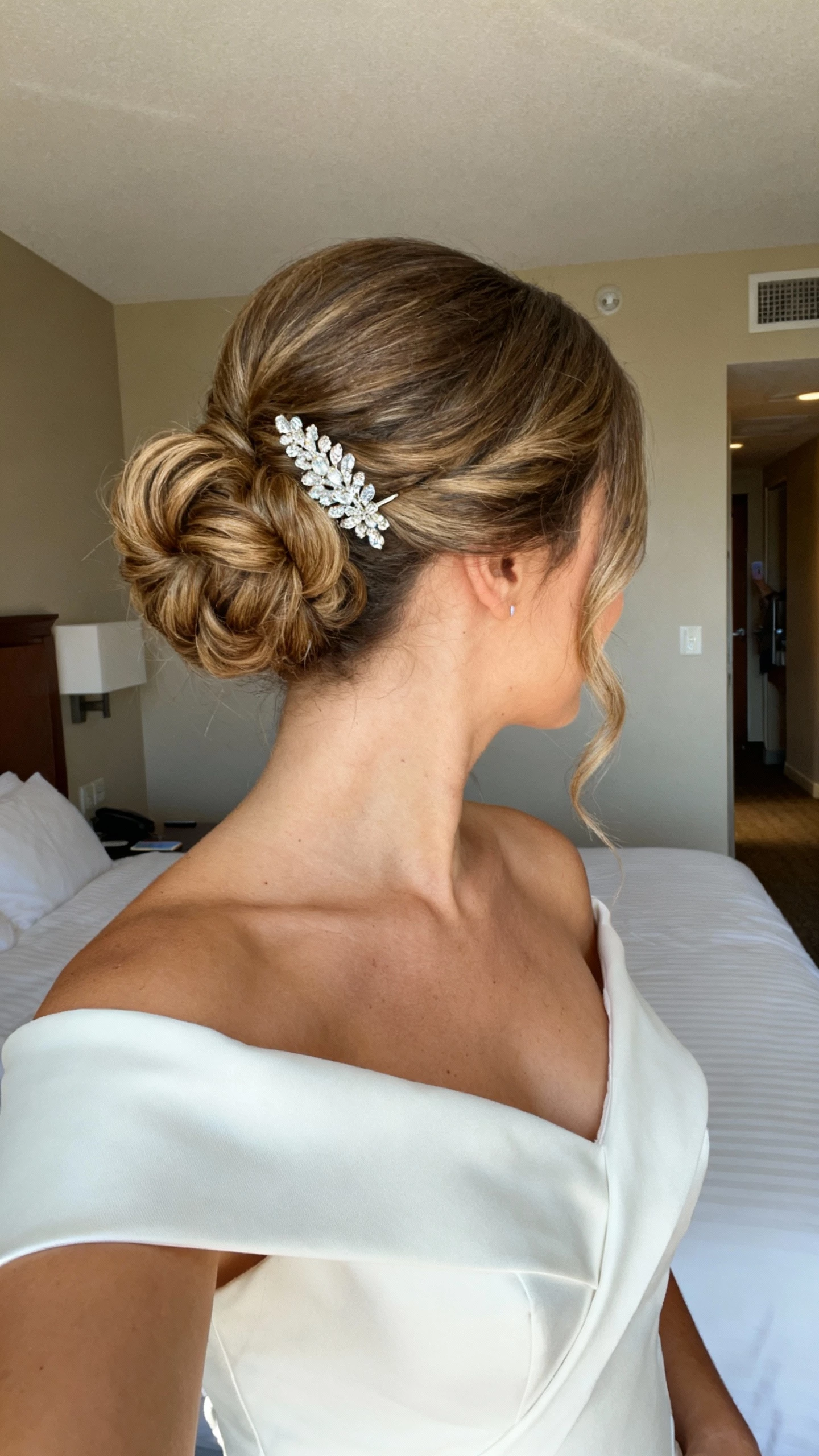 iPhone selfie from a slight back-side angle showing an elevated French twist with soft volume, crystal hair pin, structured off‑the‑shoulder bridal top; face not visible, cropped at shoulders, hotel room setting, morning light, natural iPhone quality.
