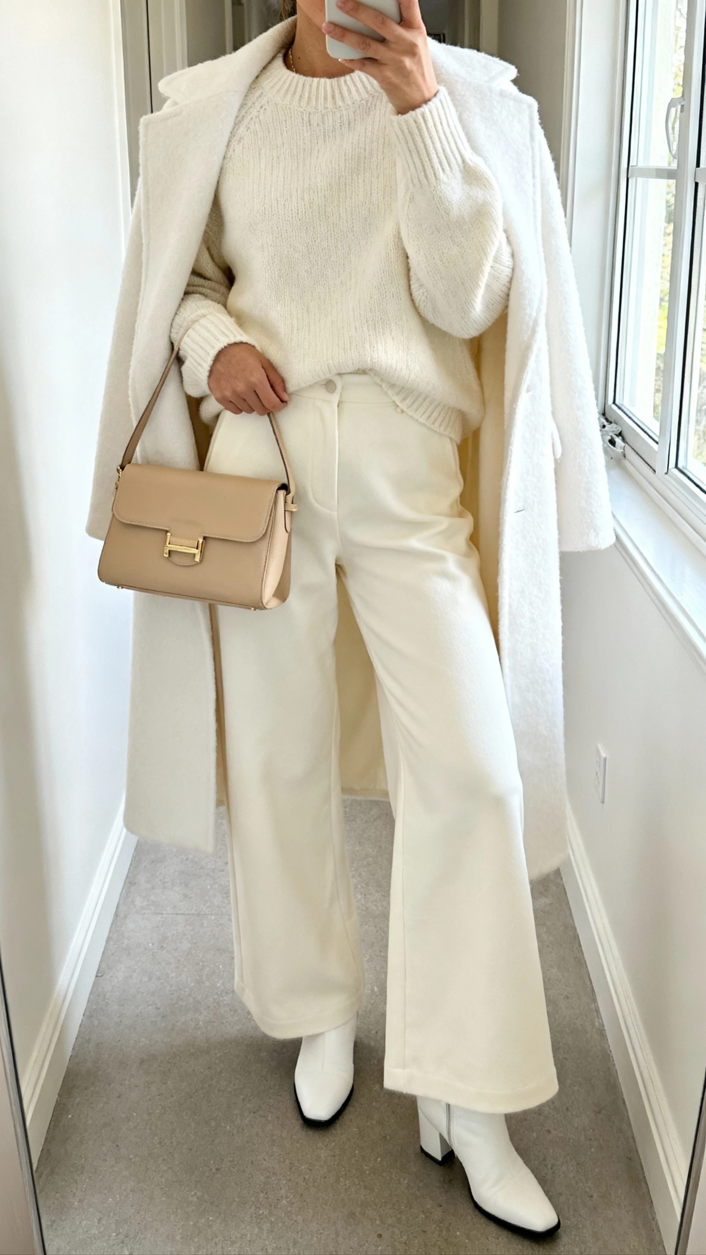 iPhone selfie from chest-down of a woman in head-to-toe winter white: cream sweater, ivory wide-leg pants, white coat, beige handbag, and white boots, face not visible, bright hallway near a window, natural daylight, casual iPhone aesthetic.