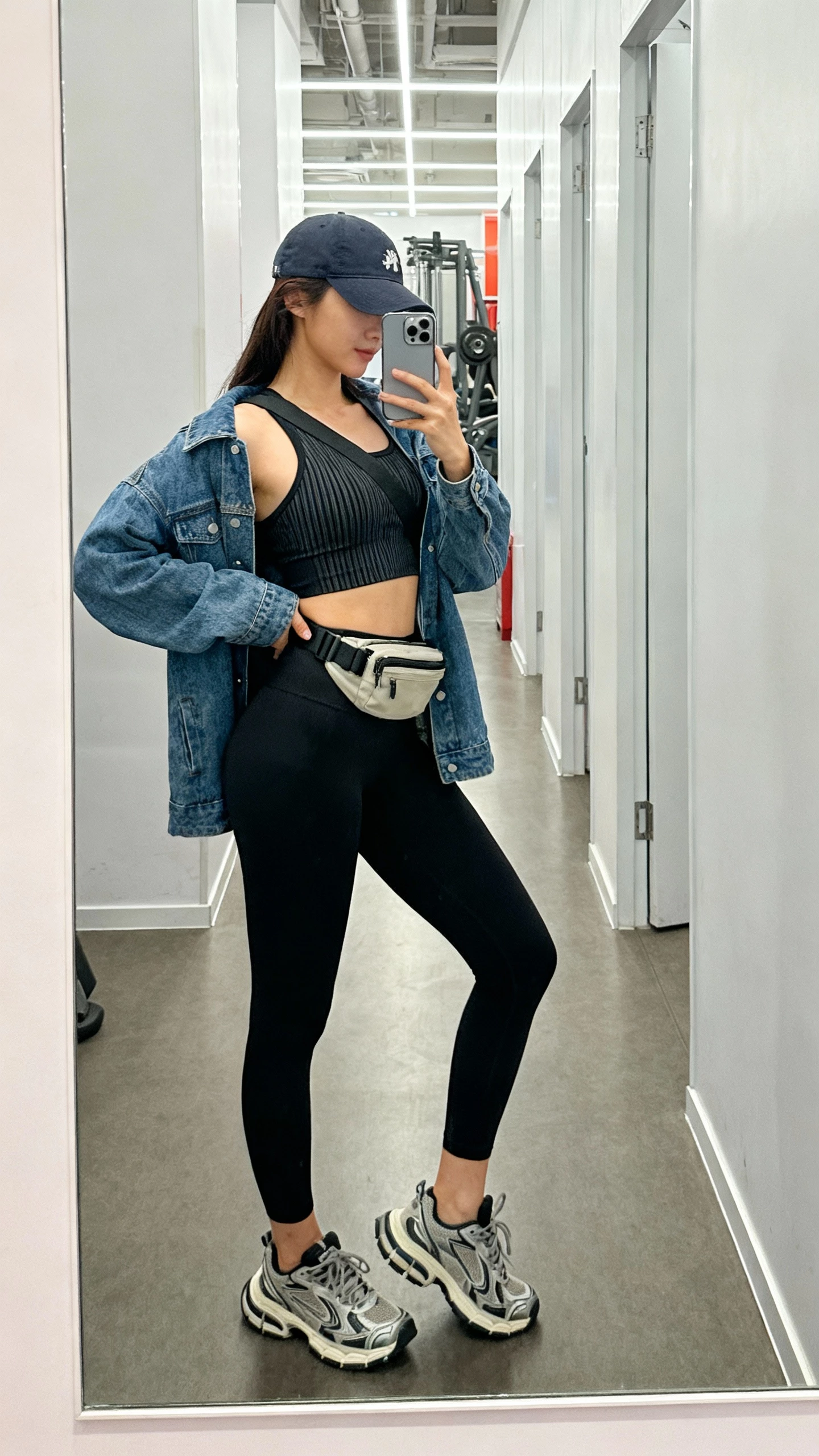 iPhone selfie side-angle of a woman in a sporty-chic athleisure look: black leggings, ribbed sports bra or fitted tank, denim jacket layered over, baseball cap, chunky trainers, crossbody belt bag, face not visible, gym hallway mirror, bright indoor lighting, natural iPhone photo quality.