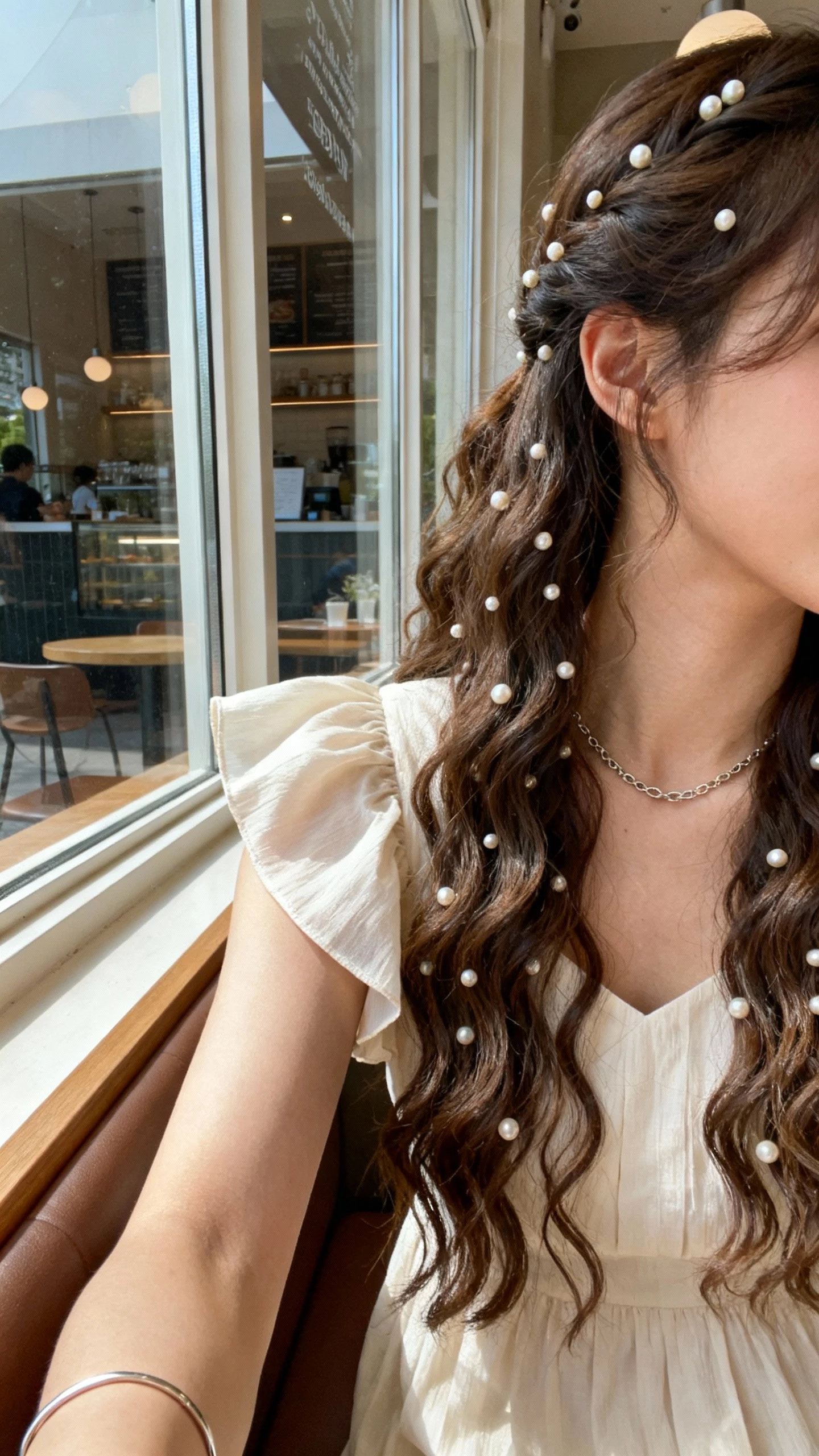 iPhone shoulder-crop selfie of a woman with a curly waterfall half-up adorned with tiny pearl accents, romantic flutter-sleeve dress, fine chain necklace, slim bracelet, face not visible, cafe window seat reflection, natural window light, natural iPhone aesthetic.