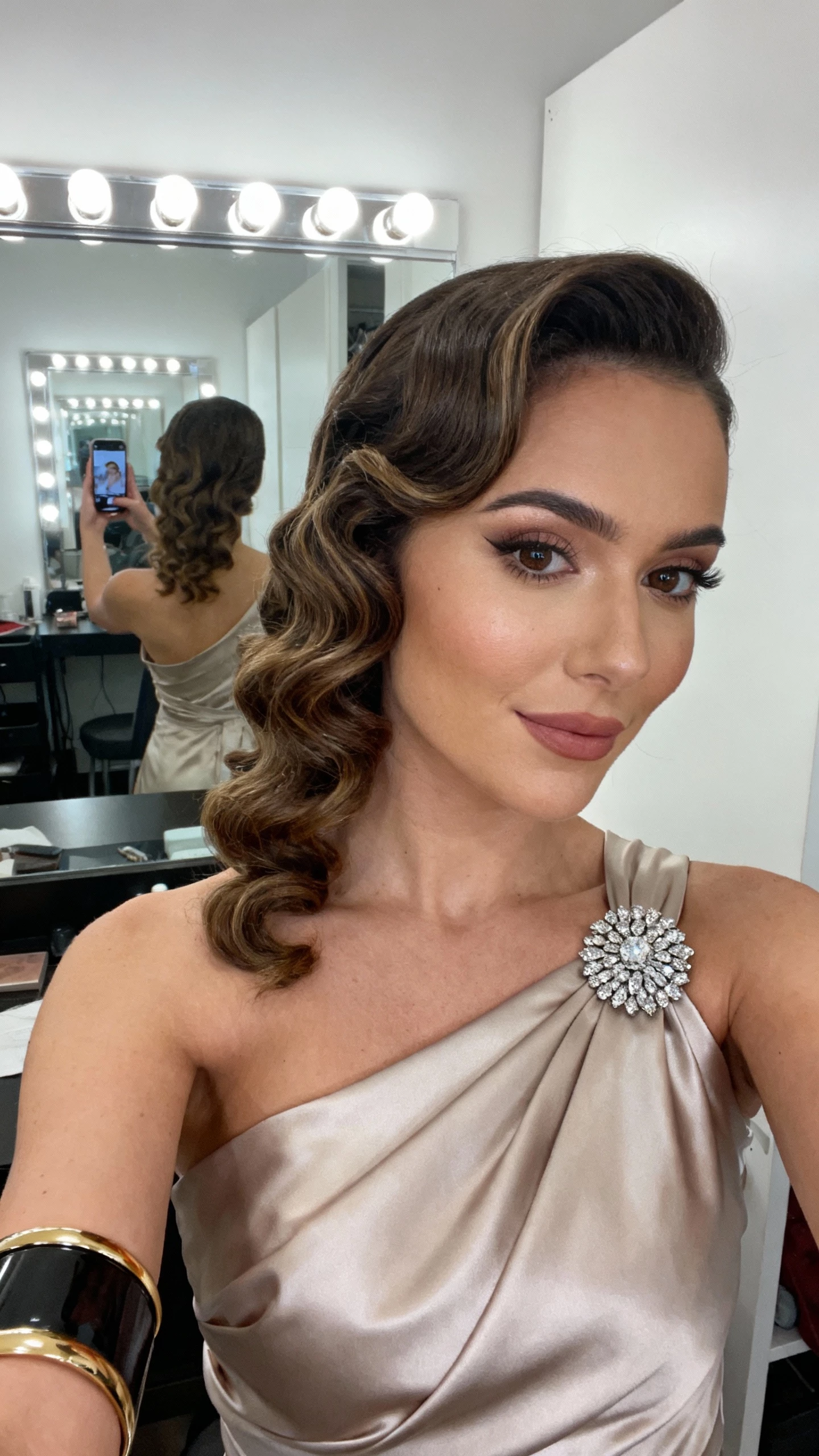 iPhone shoulder-crop selfie of a woman with side-swept curly glam waves, one-shoulder satin gown, vintage-inspired rhinestone brooch at strap, bold cuff bracelet, face not visible, dressing room mirror, soft lamp light, natural iPhone photo.