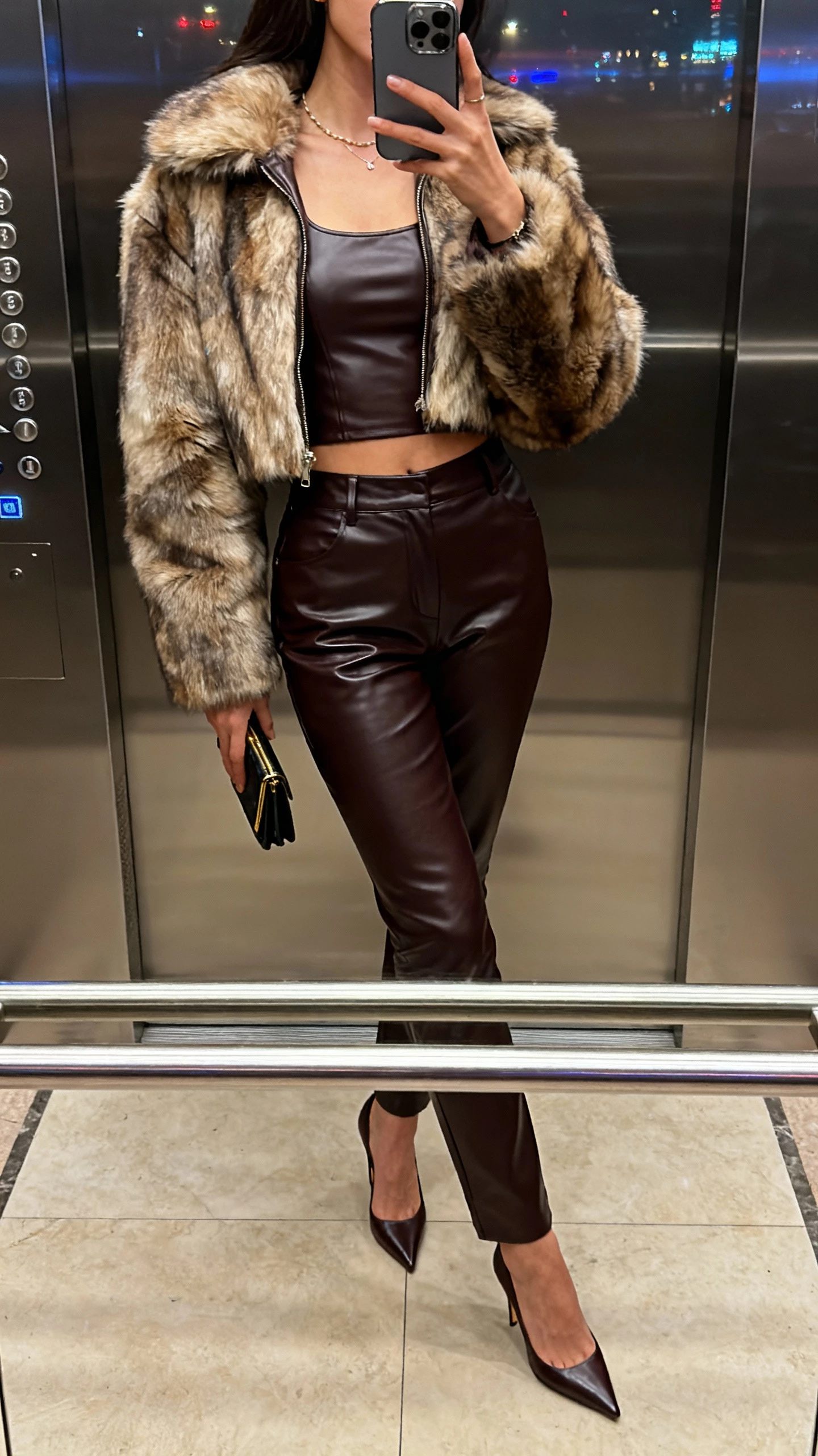 iPhone side-angle elevator selfie of a woman wearing a cropped faux-fur jacket over a fitted leather top, high-waist leather pants, slim heels, small clutch, minimal jewelry, face not visible, city-night indoor lighting, natural iPhone photo quality.