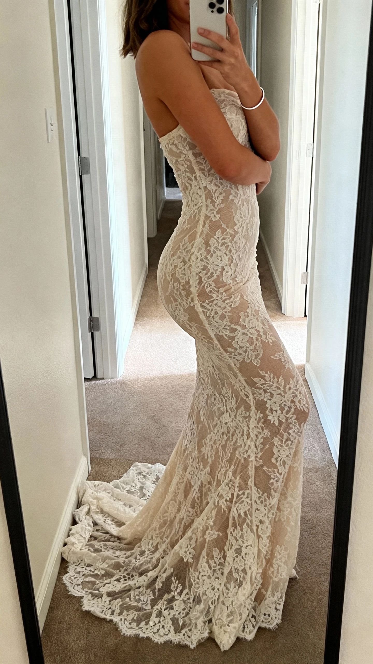 iPhone side-angle hallway mirror selfie of a woman in a sculpted mermaid lace gown that hugs the curves, subtle train, minimalist bracelet, face not visible, natural indoor daylight, casual iPhone aesthetic.