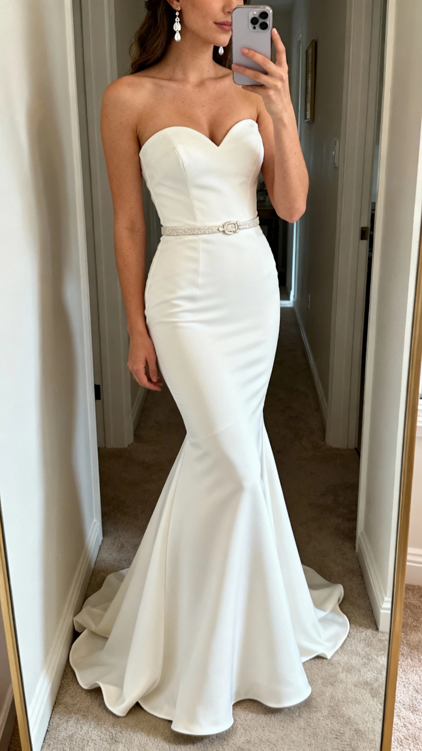 iPhone side-angle hallway mirror selfie of a woman wearing a strapless mermaid wedding dress with a soft sweetheart neckline, fitted through hips with flared hem, delicate belt, and drop earrings, face not visible, natural indoor light, casual iPhone aesthetic.