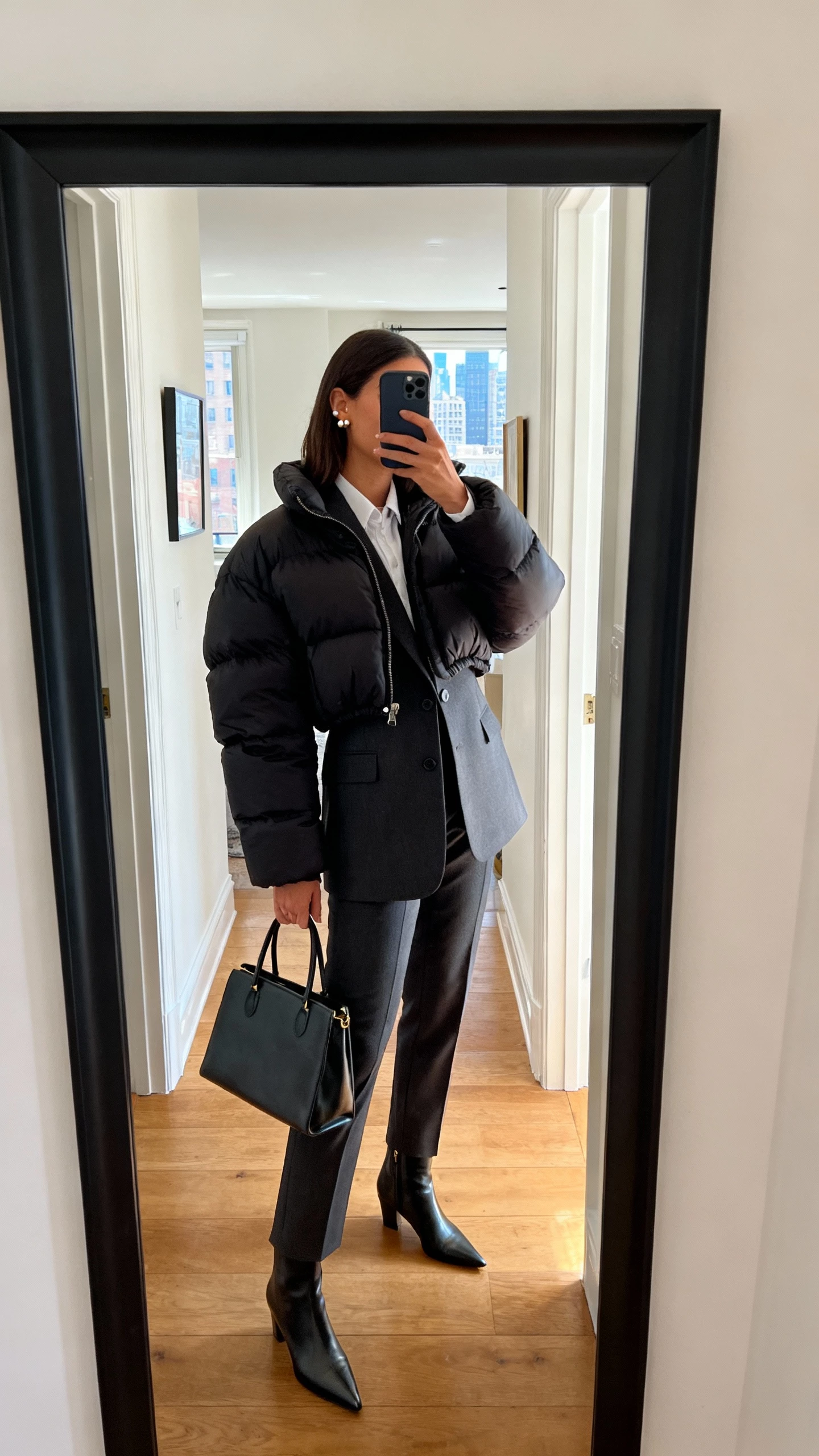 iPhone side-angle hallway mirror selfie of a woman wearing a cropped puffer jacket over a tailored blazer and crisp trousers, pointed ankle boots, structured tote, simple studs, face not visible, city apartment entryway with natural daylight, casual iPhone aesthetic