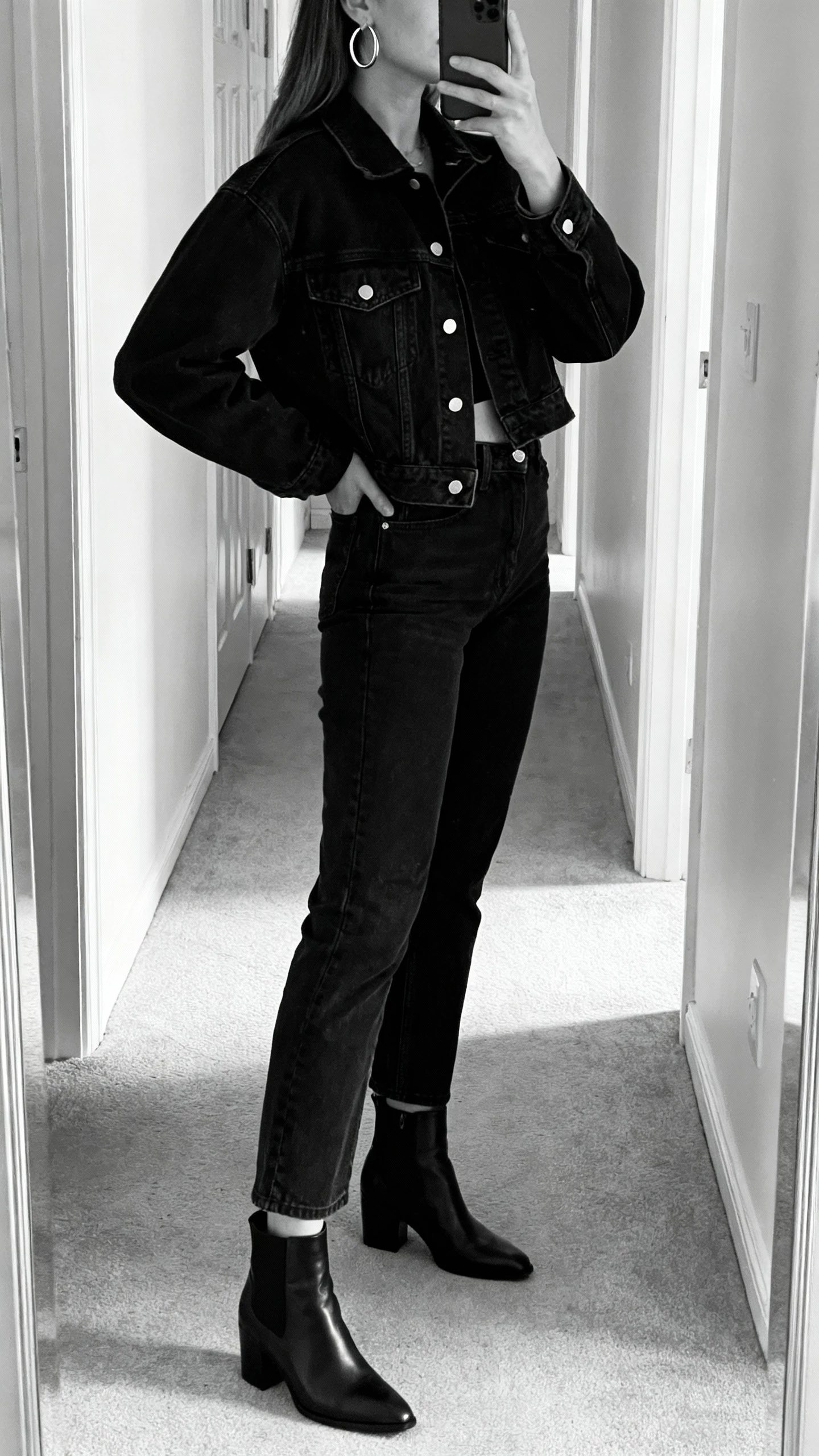 iPhone side-angle hallway mirror selfie of a woman in monochrome black denim jacket and black high-waist jeans, black ankle boots, silver hoop earrings, face not visible, indoor ambient light, natural iPhone photo quality.
