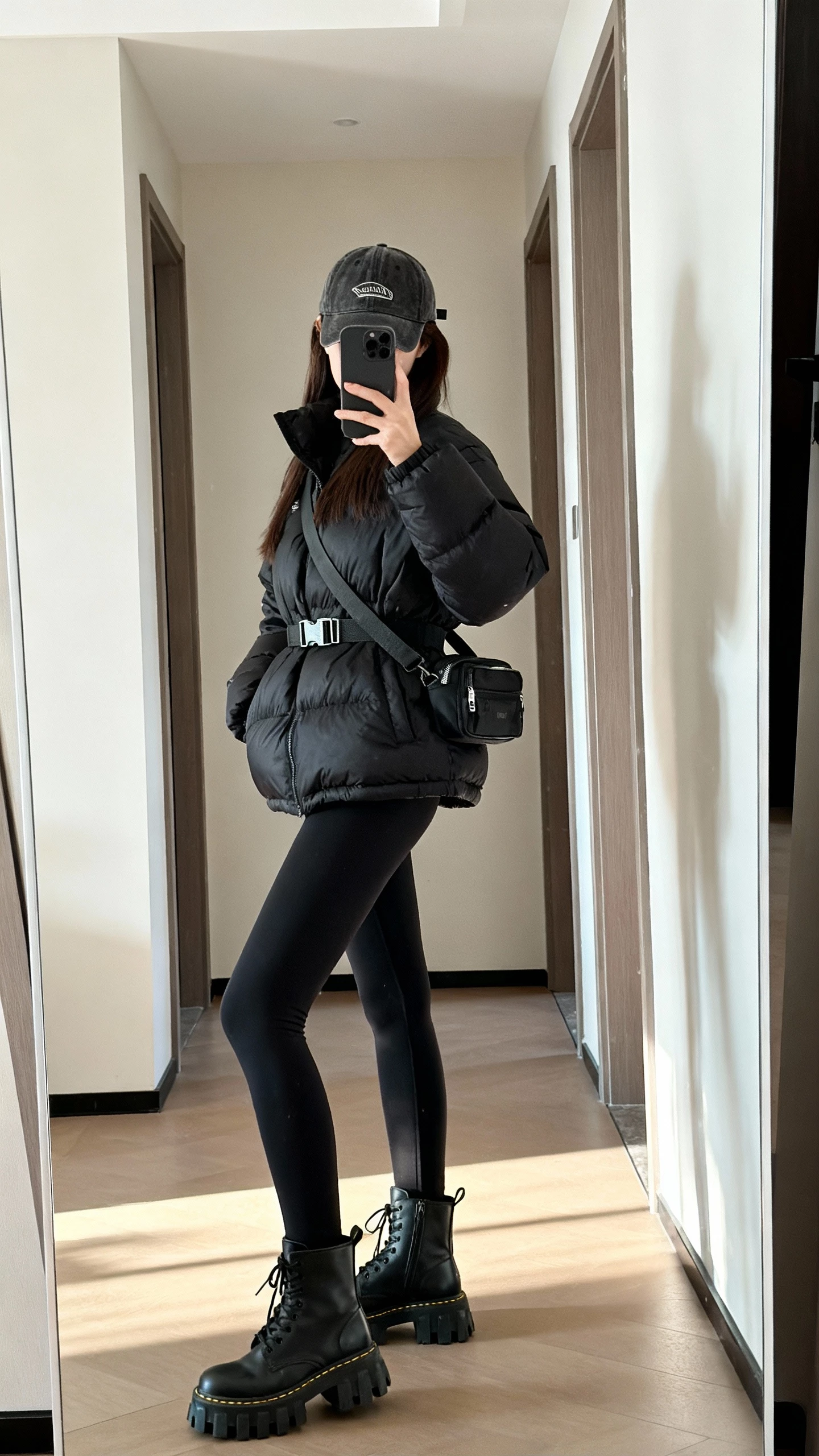 iPhone side-angle hallway mirror selfie of a woman in a black puffer jacket, matte black leggings, chunky lug-sole boots, crossbody belt bag, baseball cap, face not visible, indoor daylight, casual iPhone photo.