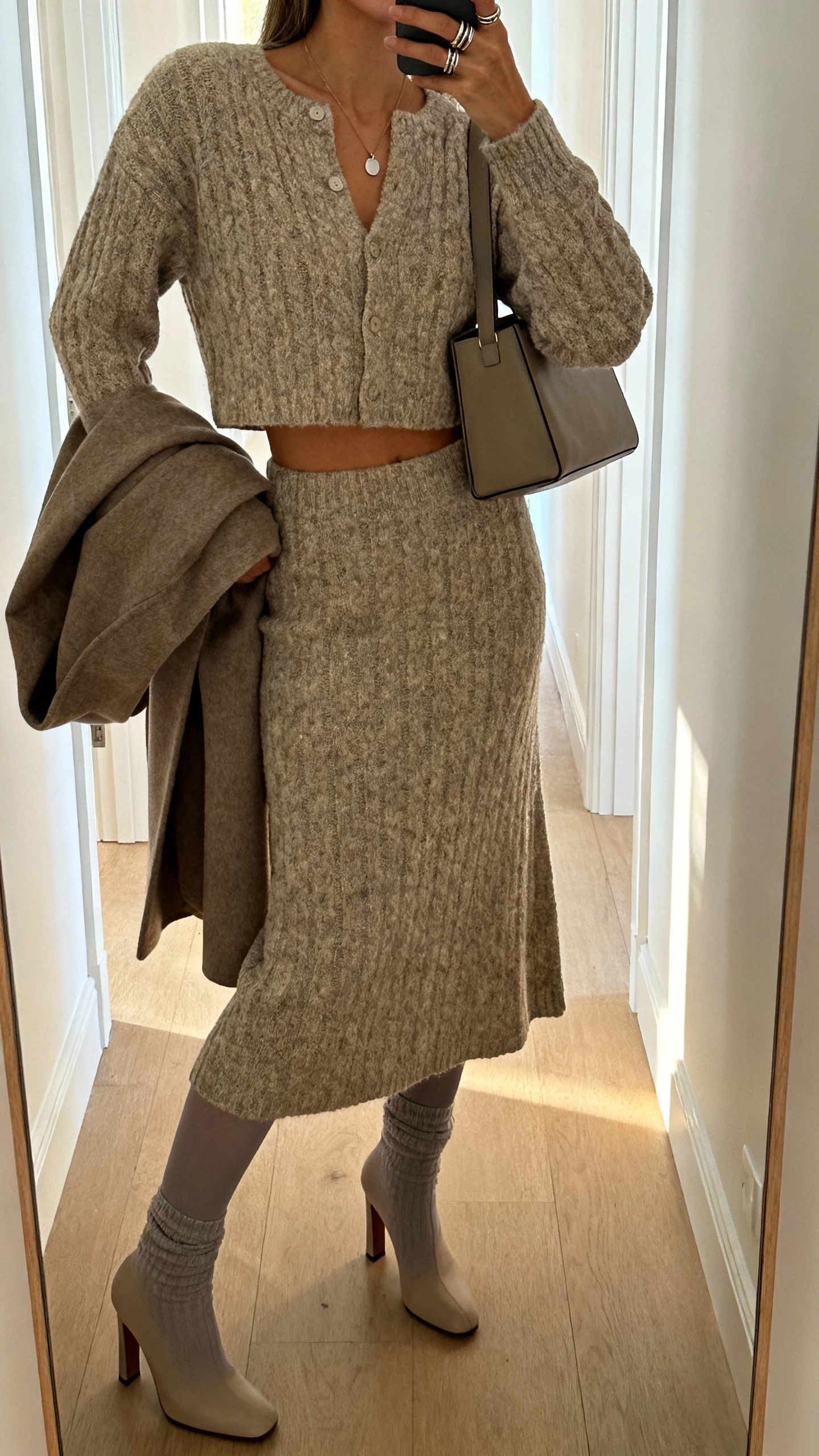 iPhone side-angle hallway mirror selfie of a woman in a knit co-ord set (cropped cardigan with matching midi skirt), heeled sock boots, minimal pendant necklace with stacked rings, boxy shoulder bag, wool wrap coat over shoulder; optional opaque tights, face not visible, morning light, natural iPhone photo quality.