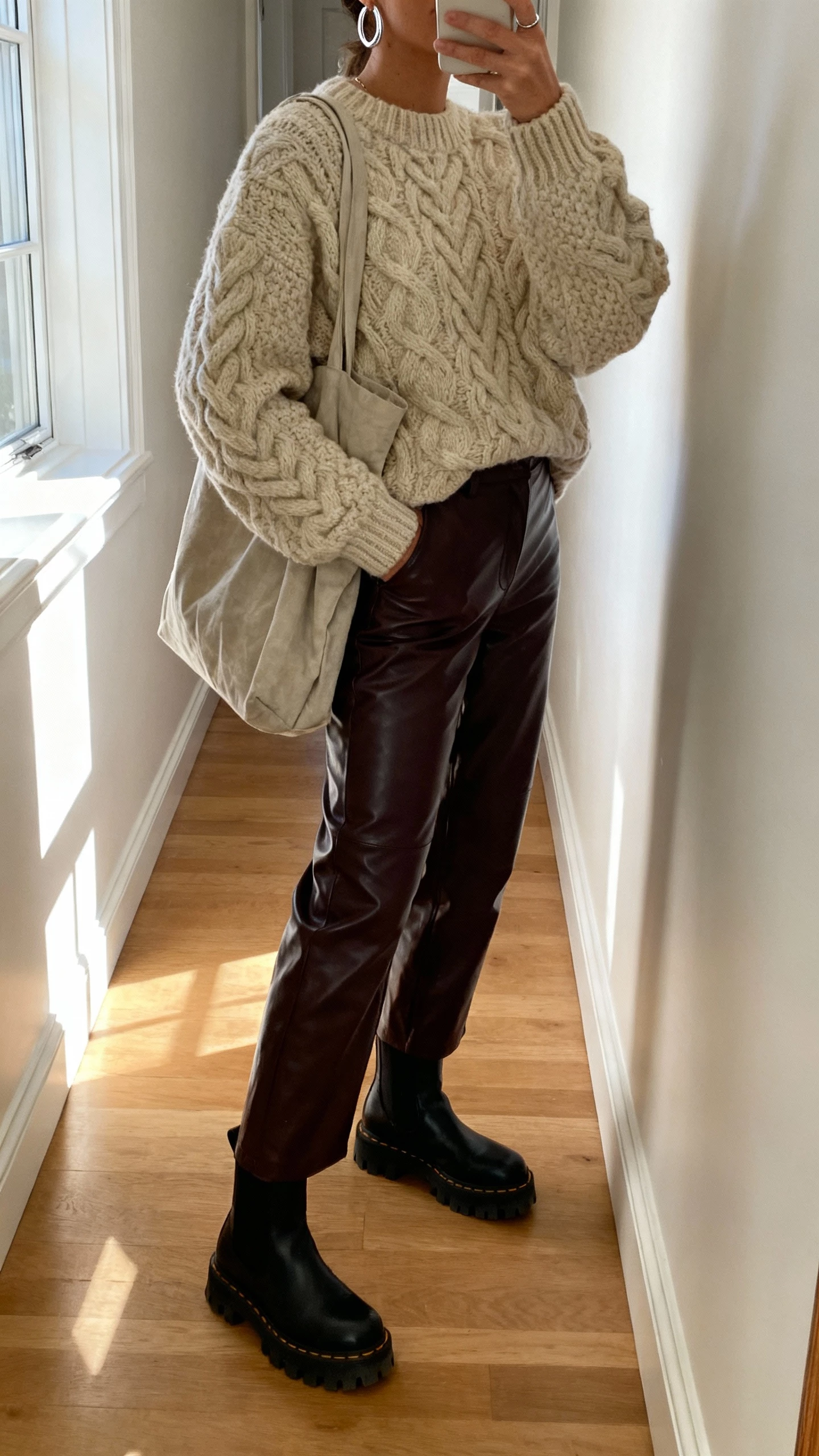 iPhone side-angle hallway selfie of a woman wearing a chunky cable-knit sweater with faux-leather pants, lug-sole ankle boots, silver hoop earrings, slouchy tote, face not visible, natural daylight from window, casual iPhone photo