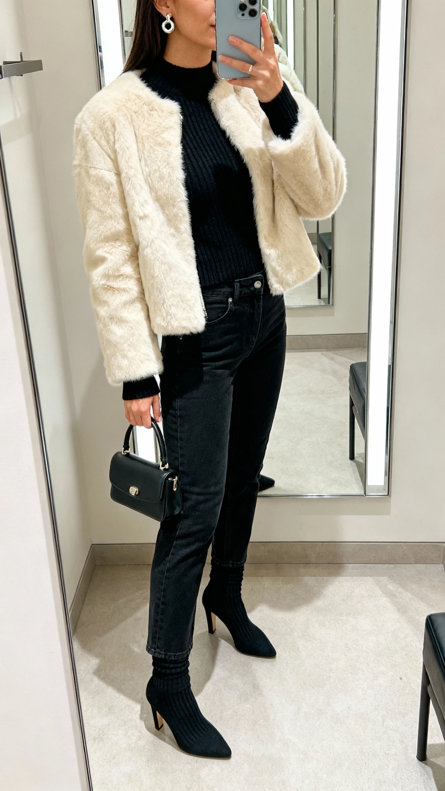 iPhone side-angle mirror selfie of a woman in a cream faux-fur hip-length jacket, fitted black crewneck knit, high-rise straight black jeans, heeled sock boots, mini top-handle bag, bold stud earrings; boutique dressing-room mirror, face not visible, soft indoor lighting, natural iPhone look.