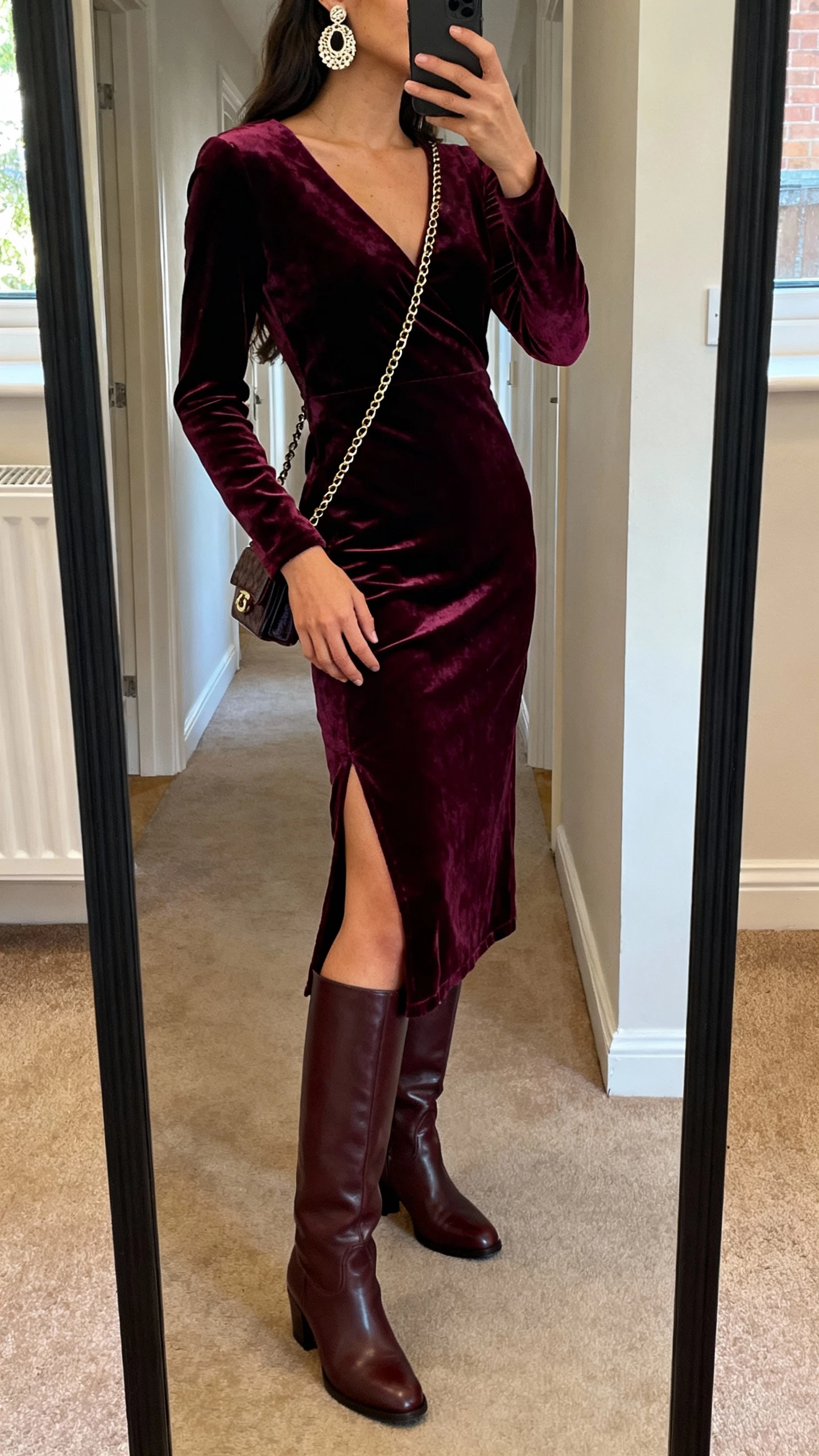 iPhone side-angle mirror selfie of a woman in a deep jewel-tone velvet midi dress with a slit, tall knee-high leather boots, statement earrings, and a small chain-strap bag, face not visible, cropped at neck, hallway mirror before going out, warm indoor lighting, natural iPhone photo quality