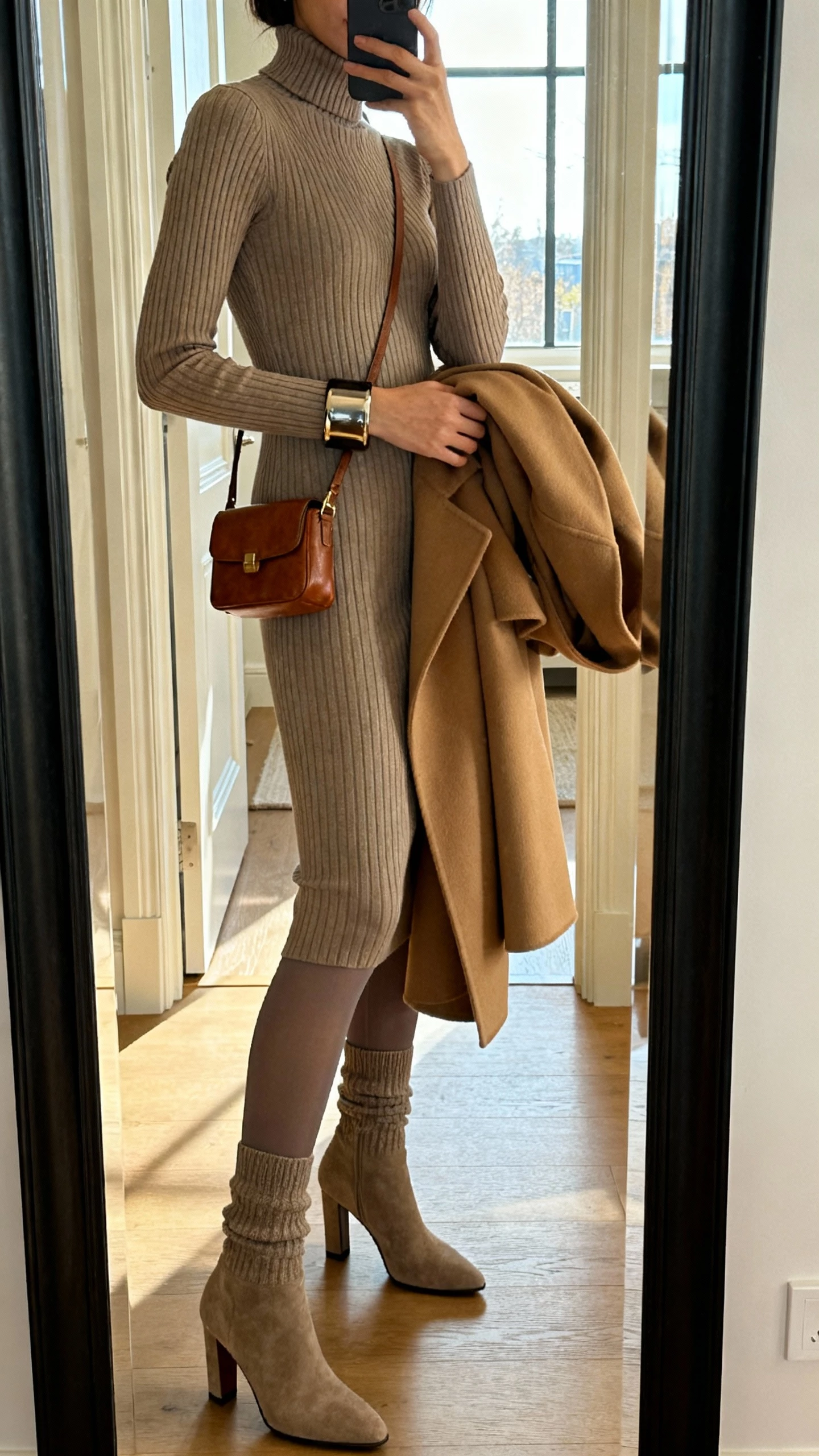 iPhone side-angle mirror selfie of a woman in a fitted ribbed knit column dress (mock neck) with opaque tights, heeled sock boots, a chunky cuff bracelet, a mini leather shoulder bag, and a structured camel coat draped over her arm; face not visible, entryway mirror, afternoon window light, casual iPhone photo.