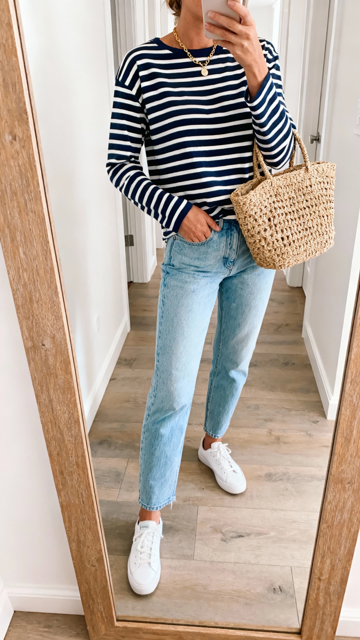 iPhone side-angle mirror selfie of a woman in a navy-and-white Breton striped top with light blue jeans, white sneakers, woven basket bag, delicate gold necklace, face not visible, hallway mirror with coastal light tones, natural daylight, casual iPhone aesthetic