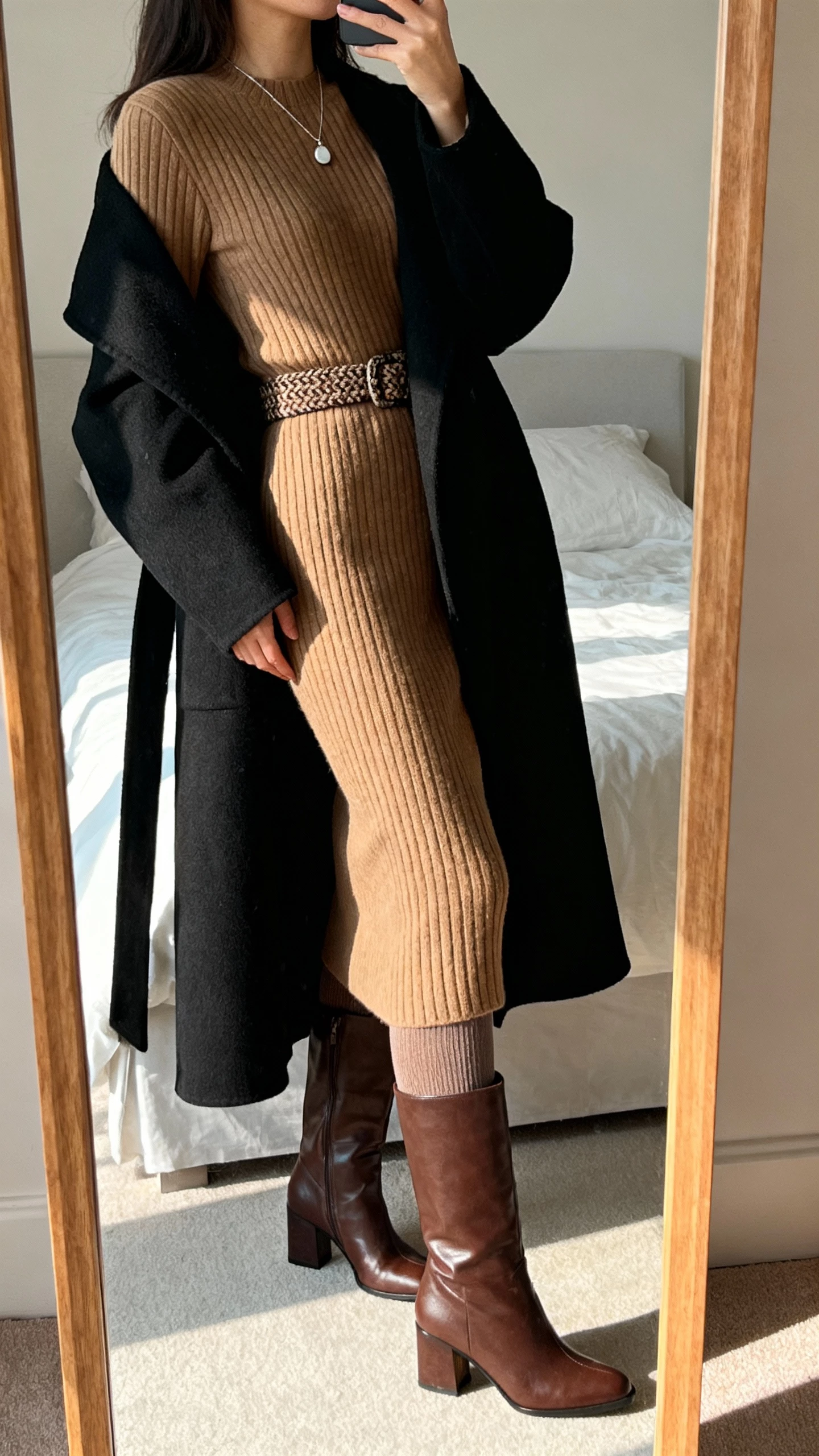 iPhone side-angle mirror selfie of a woman in a ribbed camel midi sweater dress with a textured belt, opaque thermal tights, chocolate leather knee-high block-heel boots, long black wool coat draped open, delicate pendant necklace, face not visible, bedroom mirror with soft afternoon light, natural iPhone photo quality.