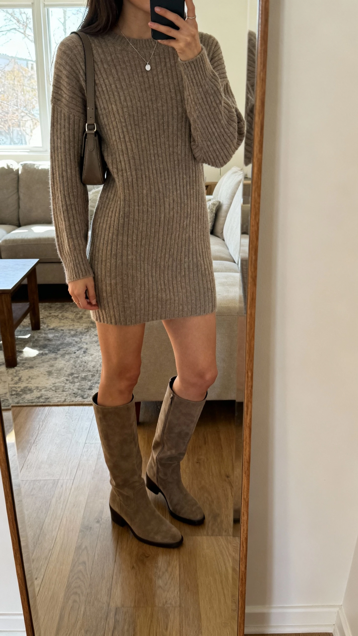 iPhone side-angle mirror selfie of a woman in a ribbed sweater dress with knee-high boots, minimal pendant necklace and crossbody bag, face not visible, living room full-length mirror, morning window light, natural iPhone photo quality.