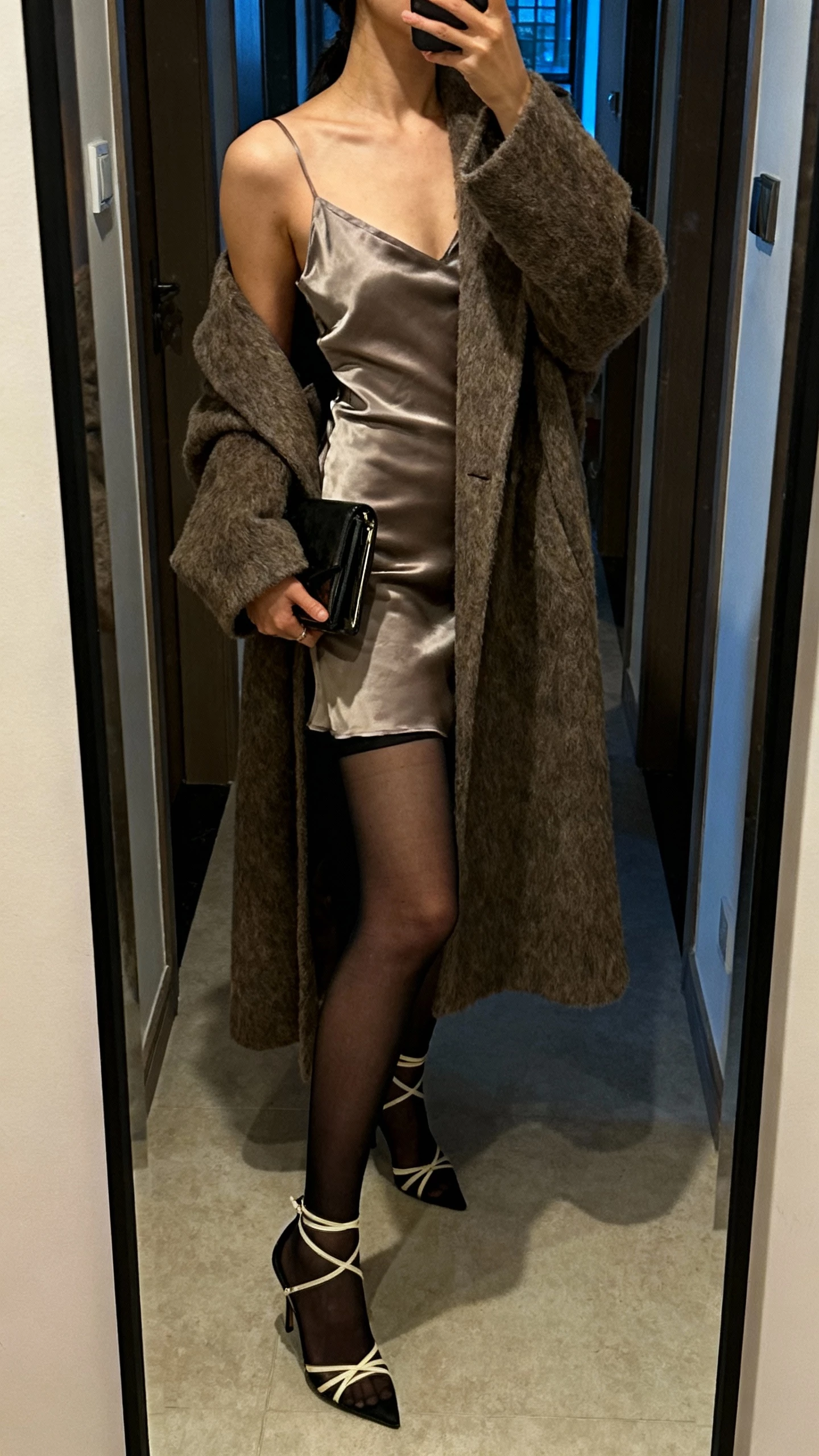 iPhone side-angle mirror selfie of a woman in a satin slip dress with sheer black tights, a long luxe wool coat draped over shoulders, strappy heels, small clutch, face not visible, hallway mirror with evening indoor lighting, natural iPhone aesthetic.
