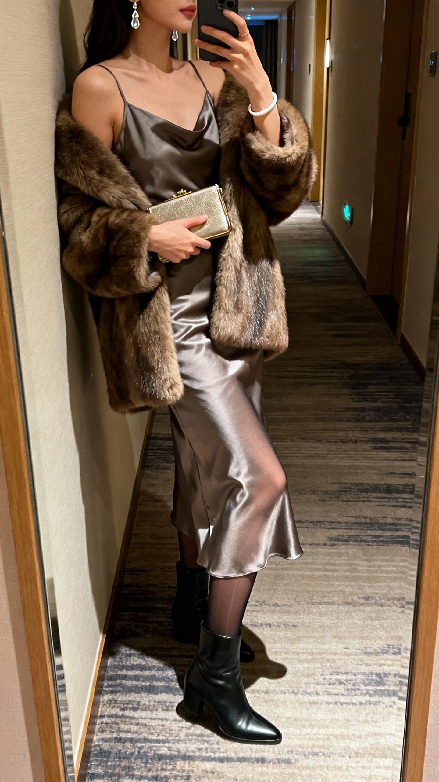 iPhone side-angle mirror selfie of a woman in a satin midi slip dress with sheer tights, sleek ankle boots, faux fur coat over shoulders, micro clutch, drop earrings and delicate cuff, face not visible, hotel hallway mirror, soft evening indoor lighting, natural iPhone photo quality.