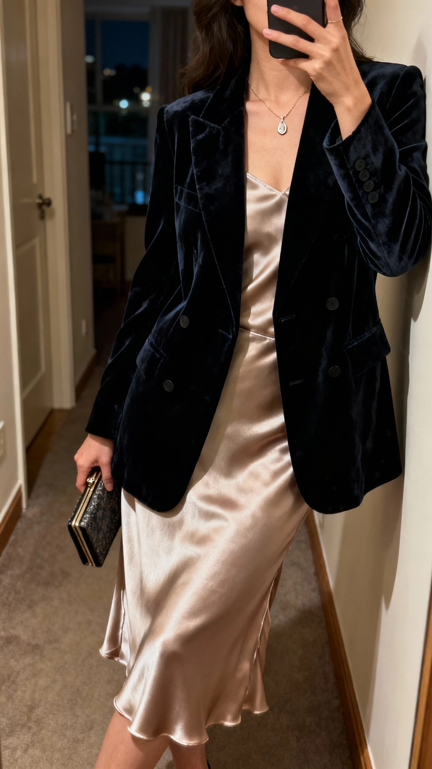 iPhone side-angle mirror selfie of a woman in a velvet blazer over a satin slip skirt, delicate pendant necklace and clutch, face not visible, hallway mirror in an apartment at night with warm indoor light, natural iPhone aesthetic.