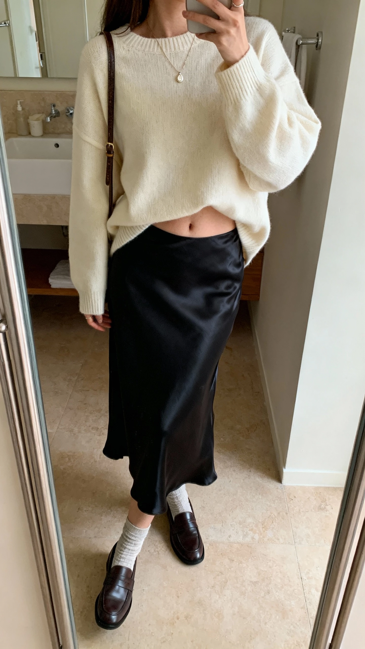 iPhone side-angle mirror selfie of a woman in an oversized cream sweater half-tucked into a black satin slip skirt, loafers with socks, delicate pendant necklace, crossbody bag, face not visible, bathroom mirror with soft indoor lighting, natural iPhone photo quality