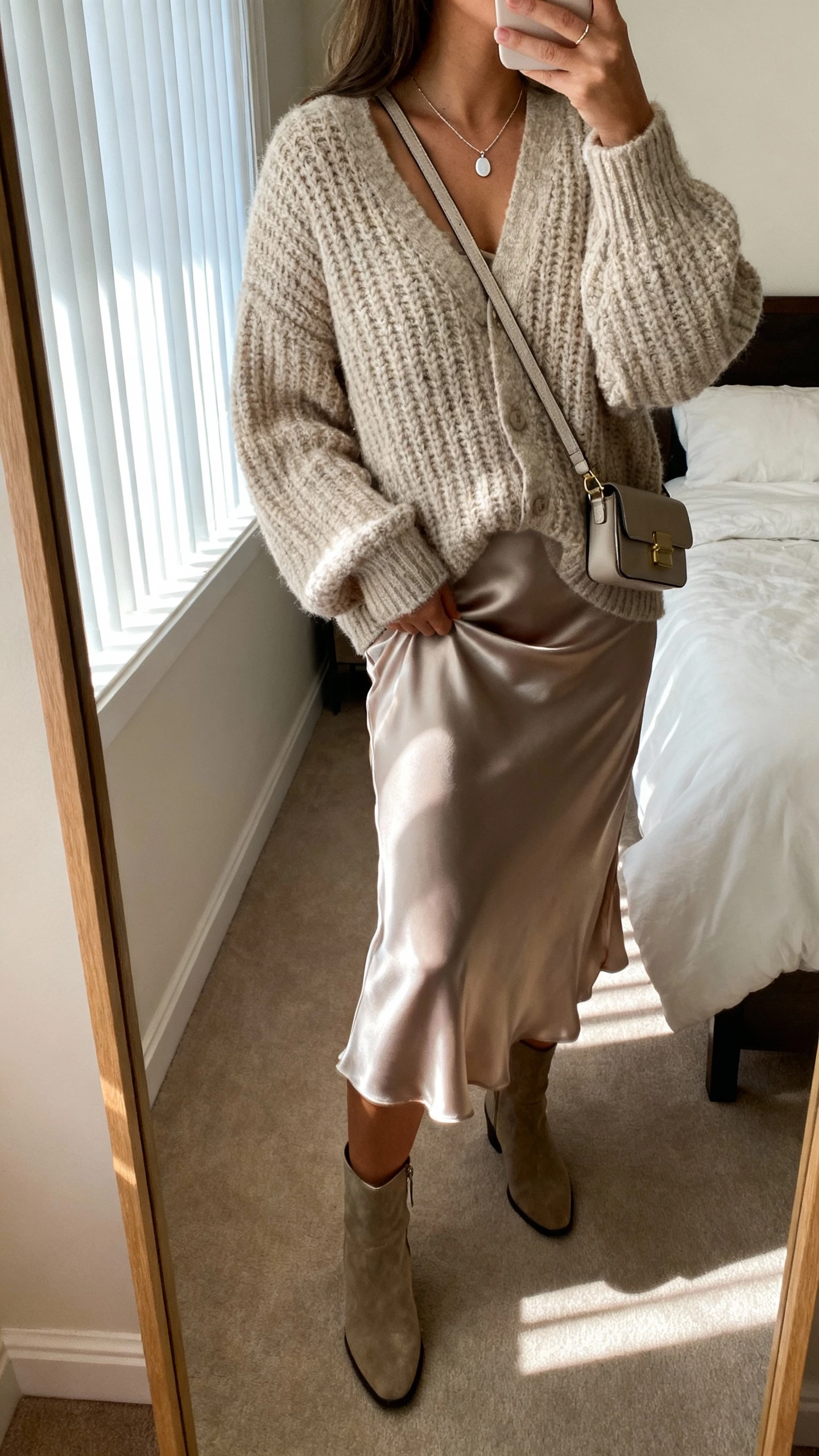 iPhone side-angle mirror selfie of a woman wearing a cozy chunky cardigan over a satin slip skirt, ankle boots, delicate pendant necklace, small crossbody bag, face not visible, bedroom full-length mirror, soft afternoon window light, natural iPhone aesthetic
