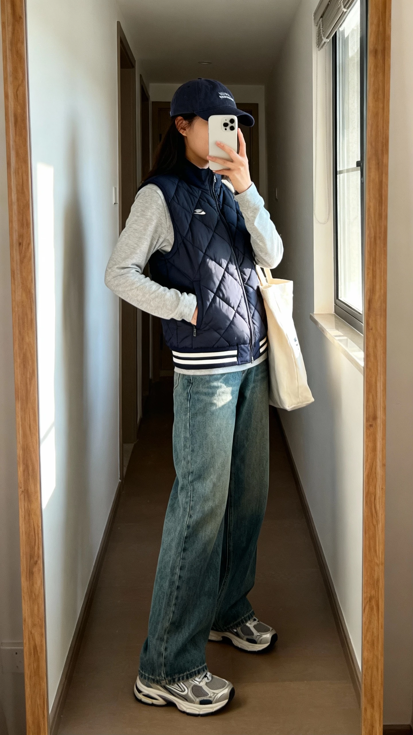 iPhone side-angle mirror selfie of a woman wearing a sporty quilted vest over a long-sleeve tee, wide-leg jeans, dad sneakers, baseball cap, tote bag, face not visible, hallway mirror with window light, natural iPhone photo quality.