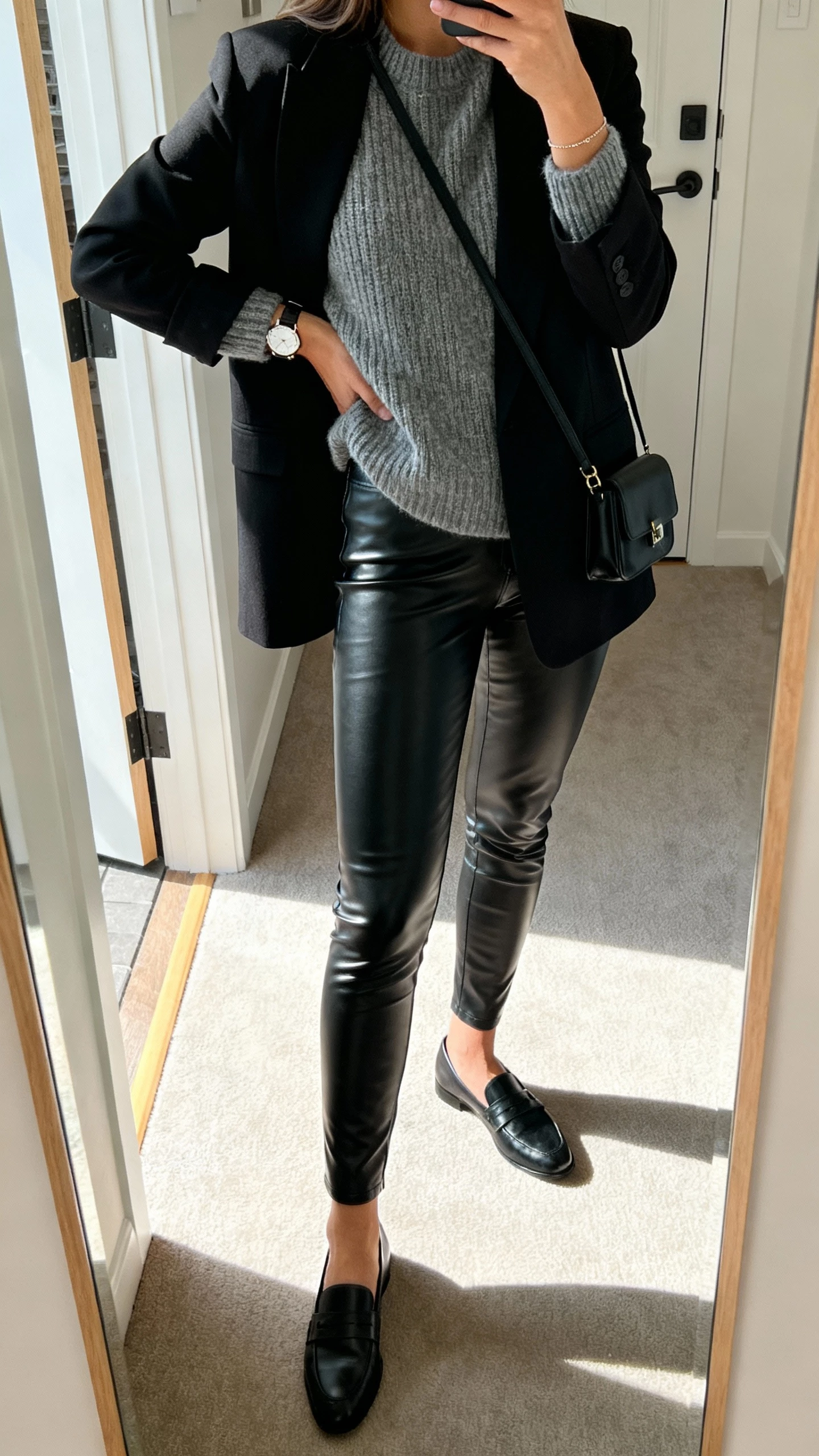 iPhone side-angle mirror selfie of a woman wearing leather-look black leggings, a polished gray knit sweater, sharp black blazer, black loafers, small black crossbody, delicate watch, face not visible, apartment entryway mirror with daylight, natural iPhone aesthetic.
