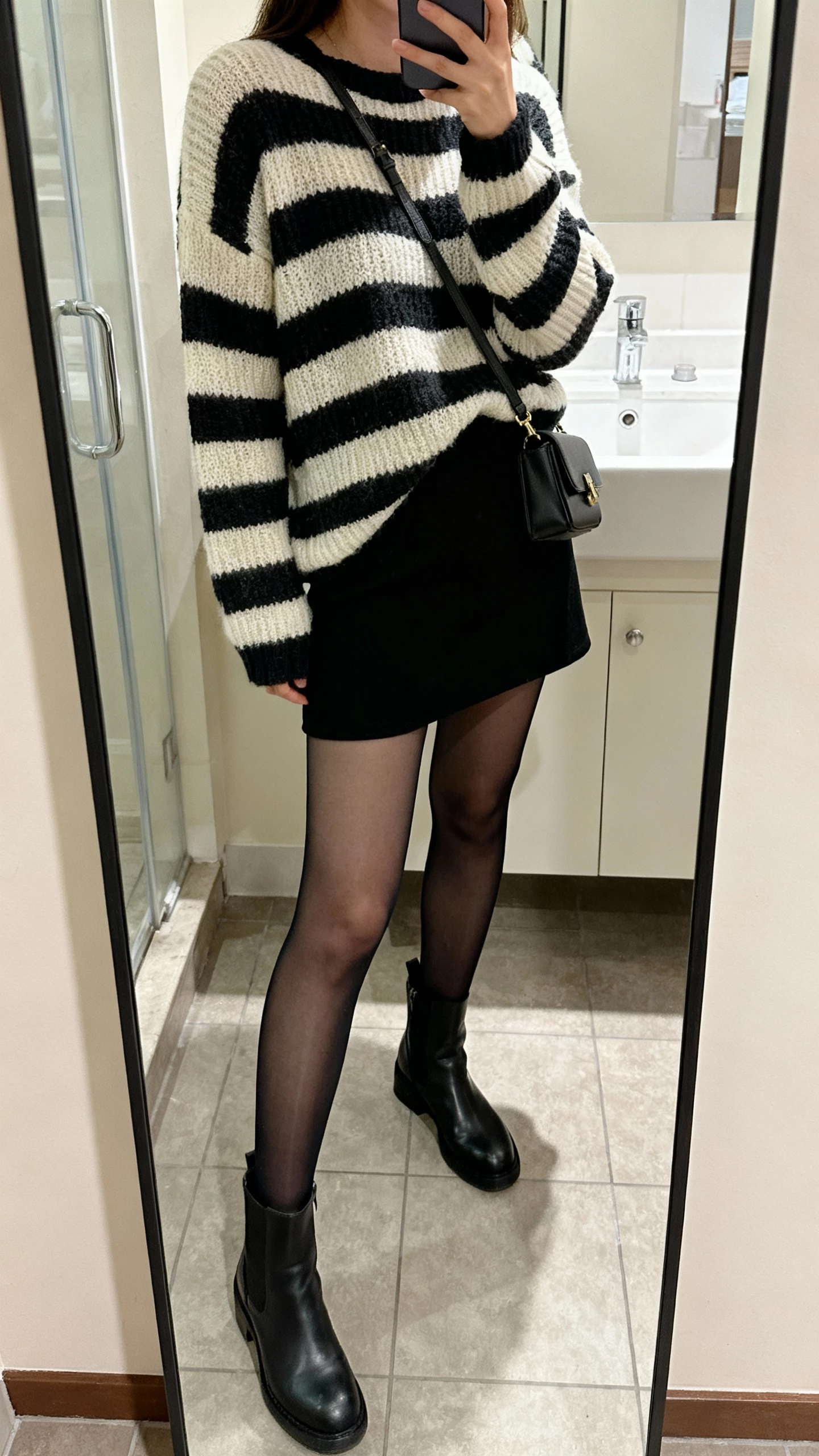 iPhone side-angle mirror selfie of a woman wearing a striped knit sweater, black mini skirt, sheer black tights, and ankle boots with a small crossbody, face not visible, bathroom mirror, soft indoor lighting, natural iPhone photo quality.